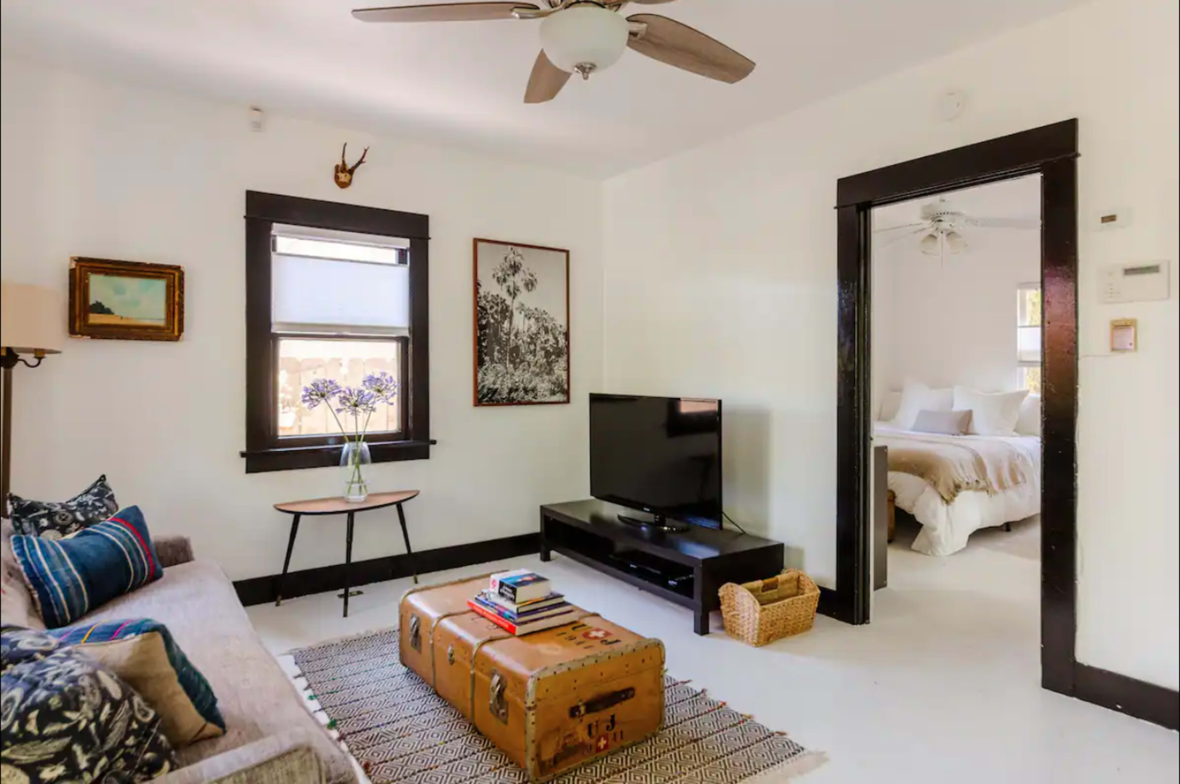 The image shows a living room with a sofa, a coffee table with stacked books, a television on a stand, and a doorway leading into a bedroom with a bed visible.