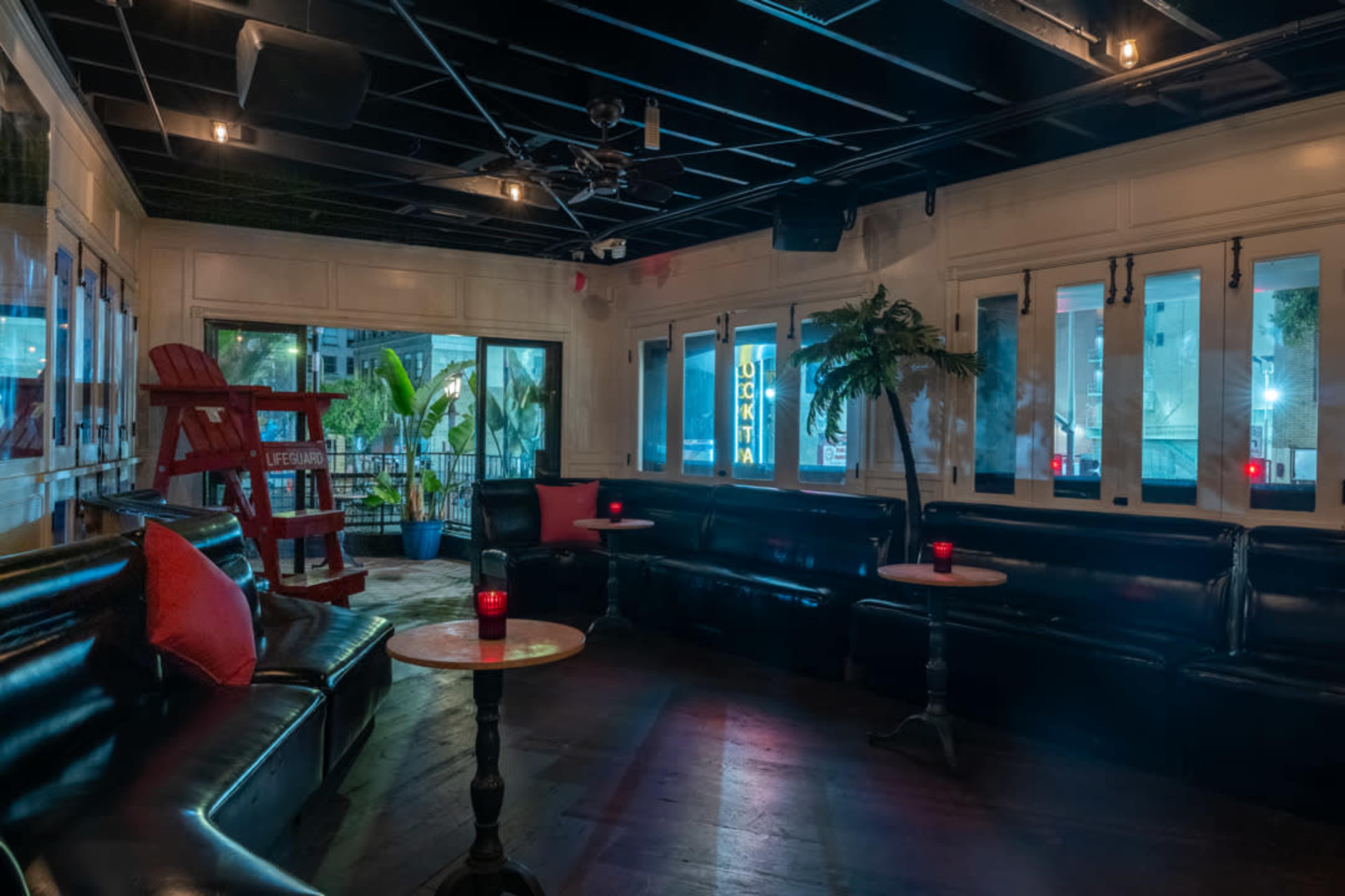 The image shows a dimly lit lounge area with black leather couches, small tables, and large windows showcasing a tropical setting outside.