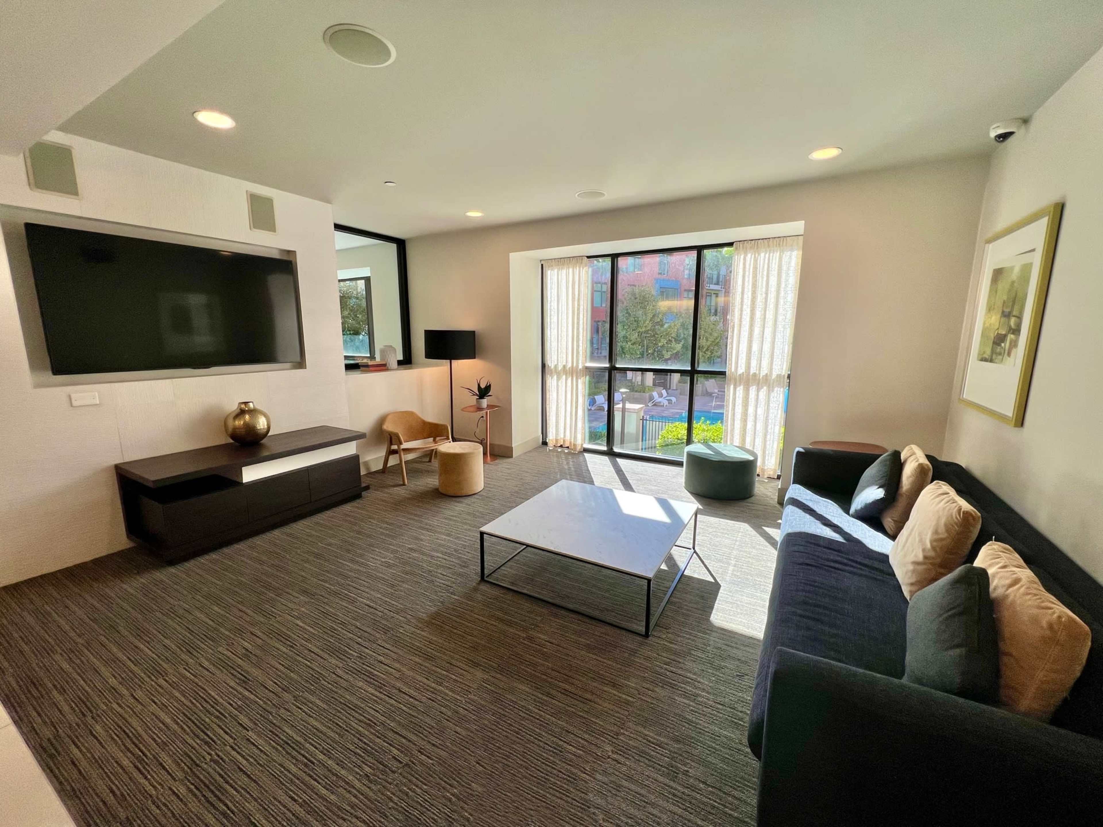 A modern living room with a large window, a dark sofa, a coffee table, a lamp, and a television mounted on the wall.