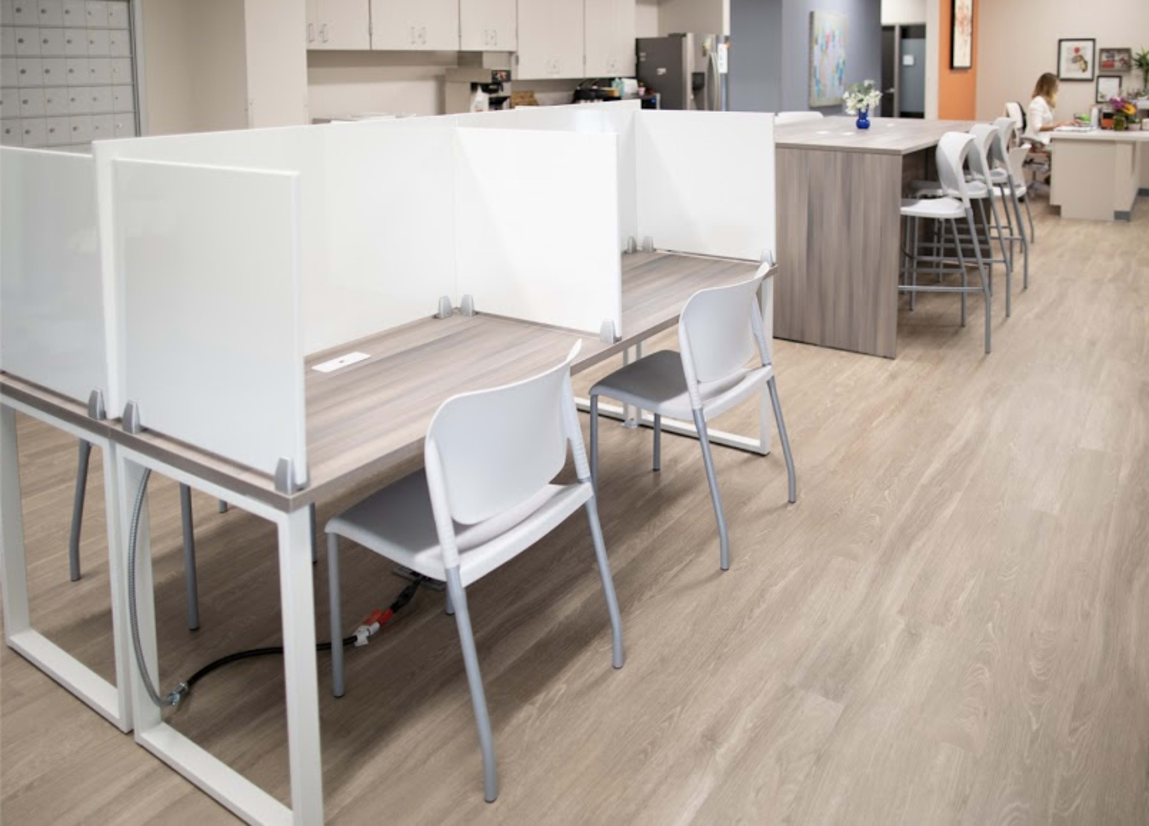 A modern workspace with dividers, featuring several empty desks and chairs, alongside a kitchen area in the background.