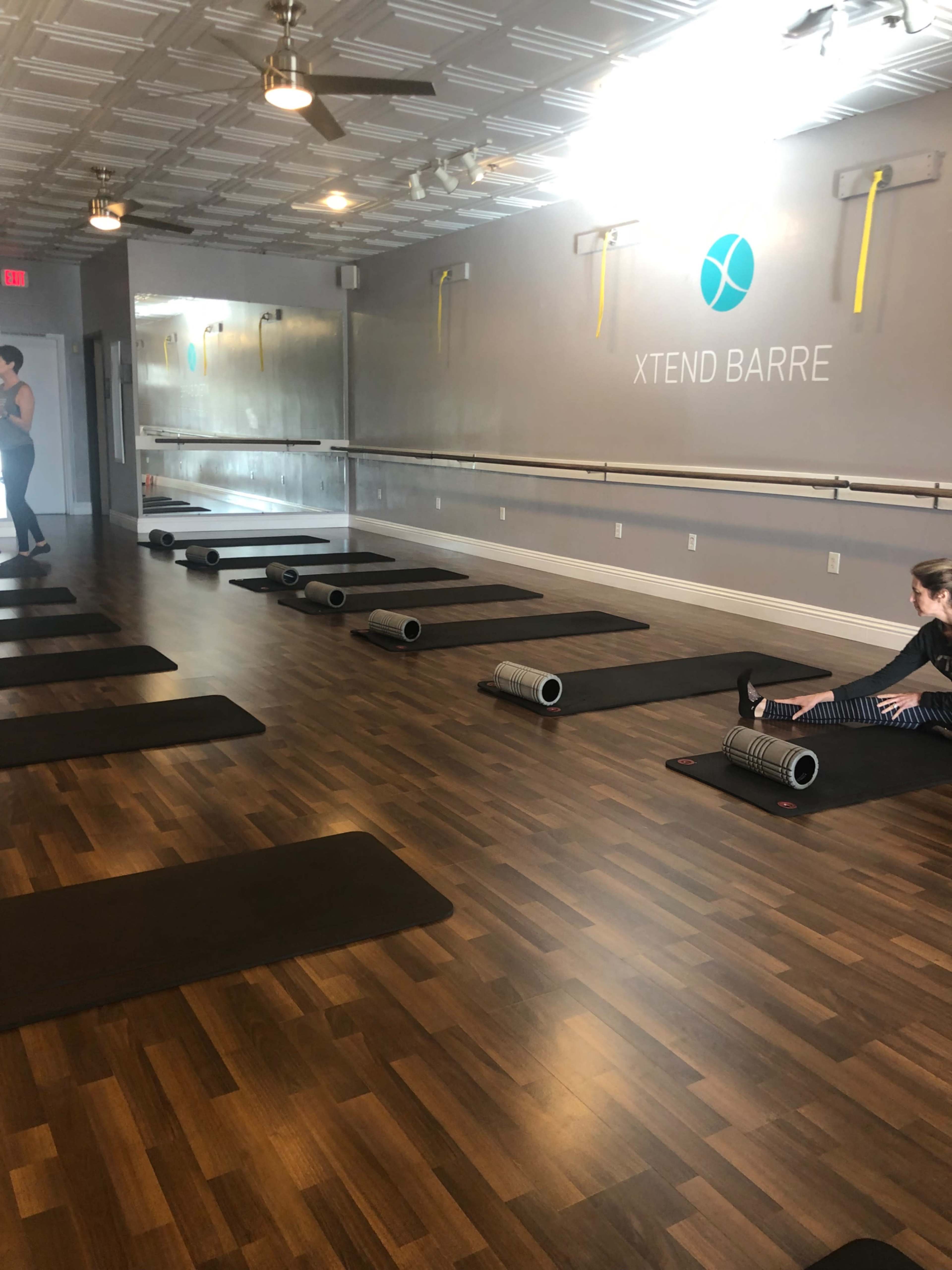 The image shows a yoga or fitness studio with several exercise mats lined up on a wooden floor, and individuals preparing for a workout near the wall with "XTEND BARRE" signage.