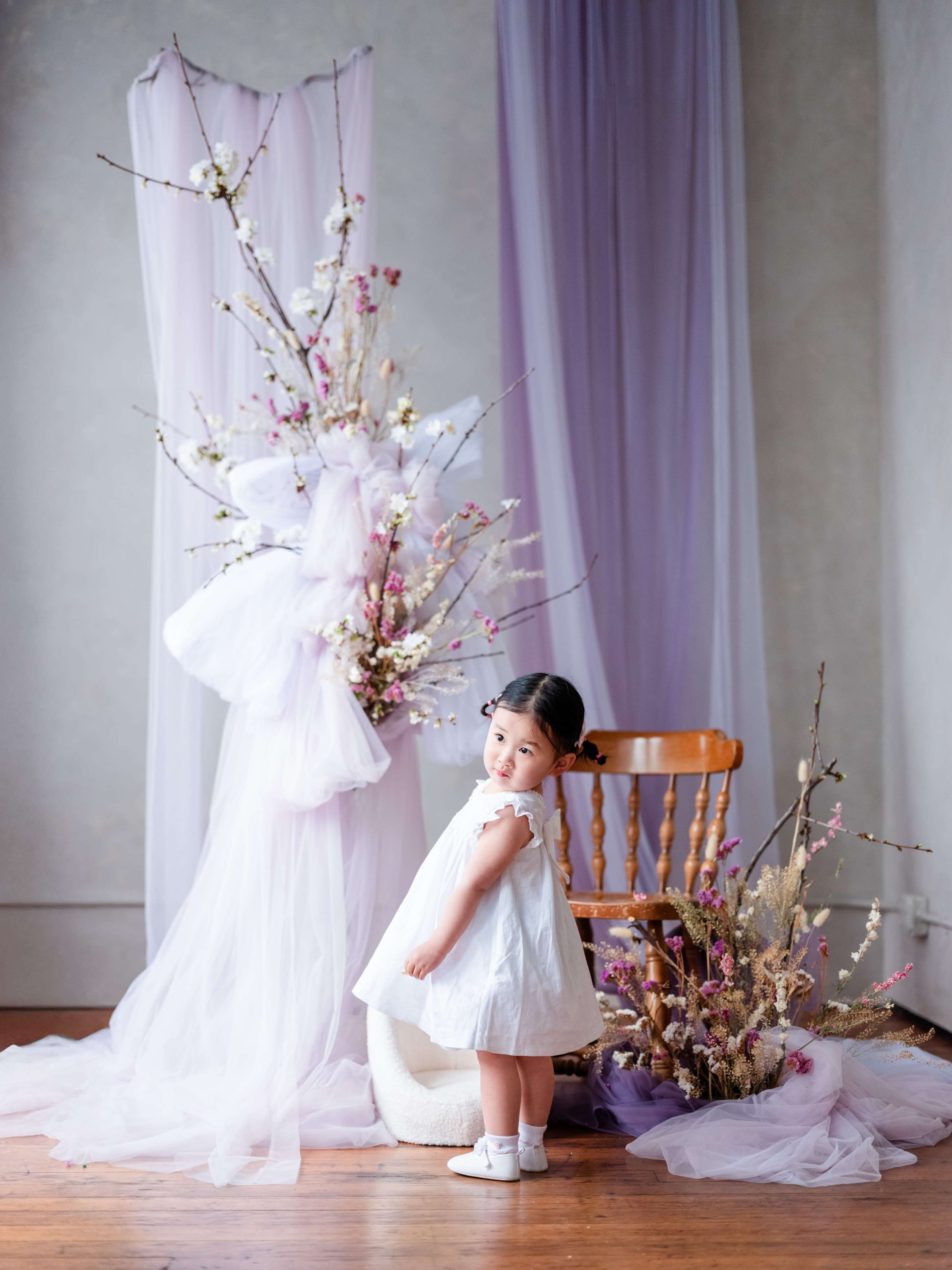A young girl in a white dress stands beside a wooden chair and a decorative floral arrangement.