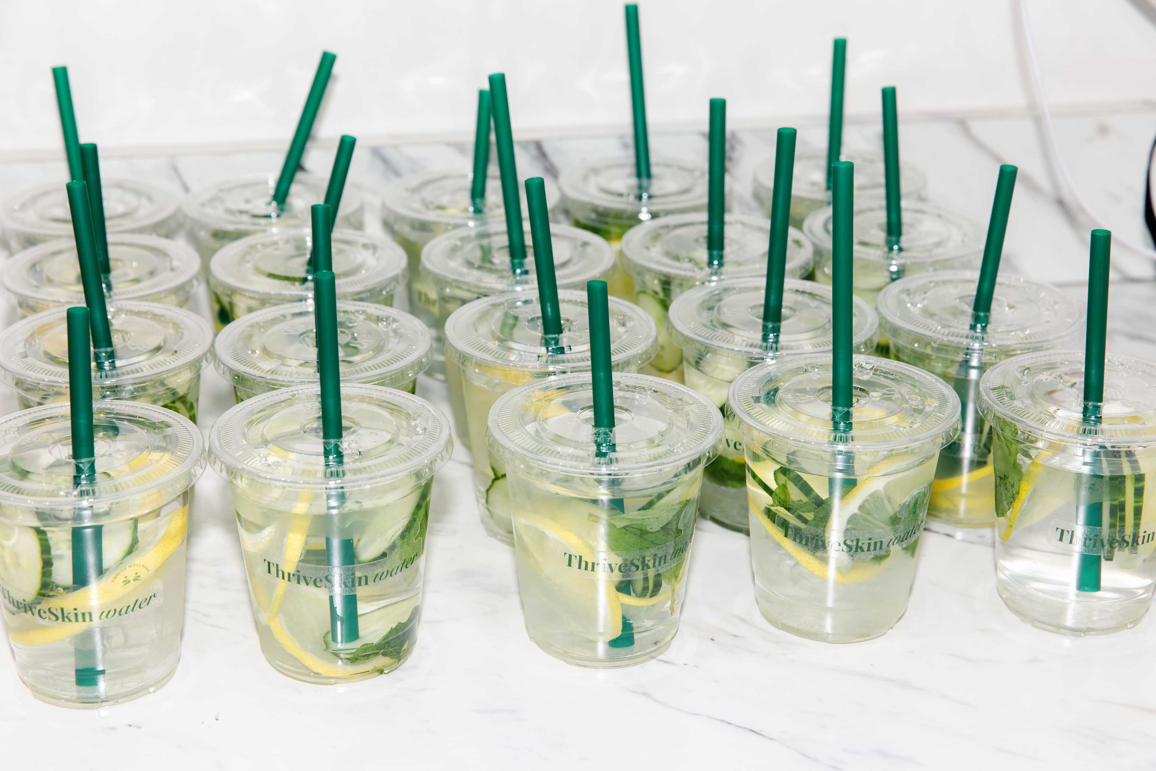 The image shows numerous clear plastic cups filled with water, lemon slices, and green straws arranged neatly on a countertop.