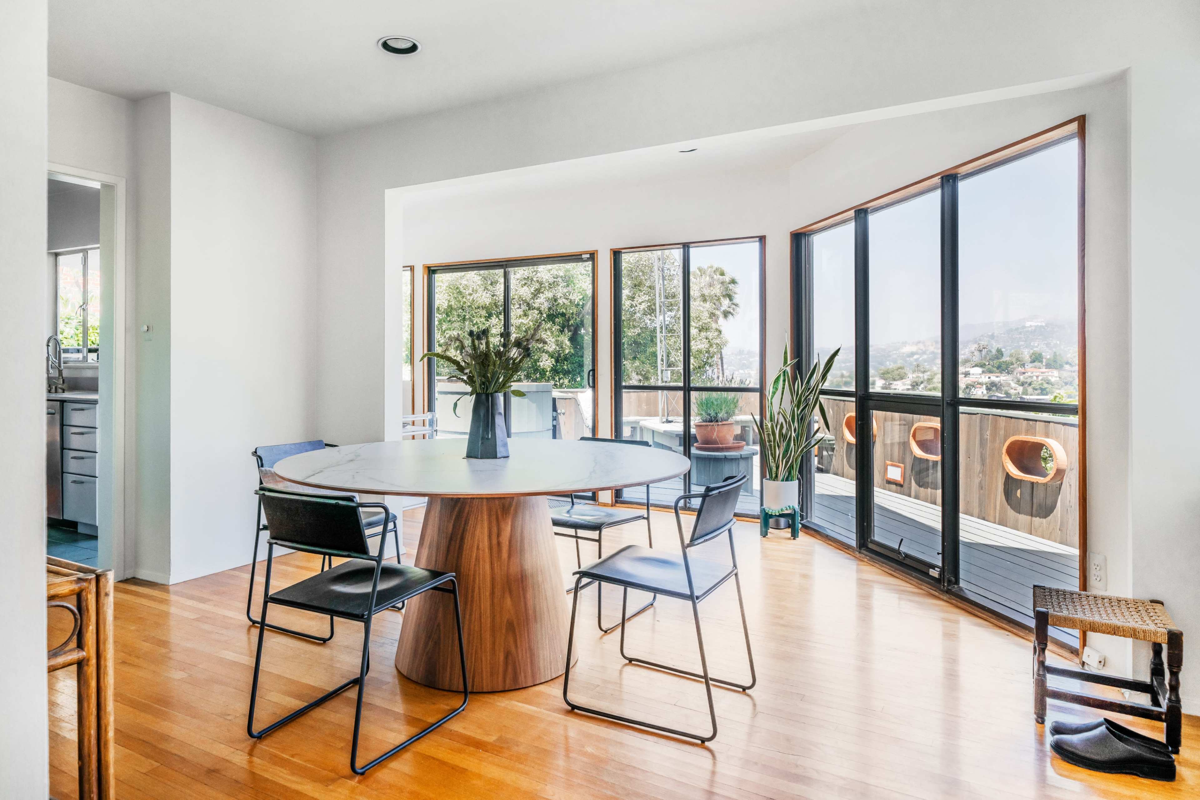 A modern dining area features a round wooden table surrounded by metal chairs, large windows showcasing a view, and wooden flooring.