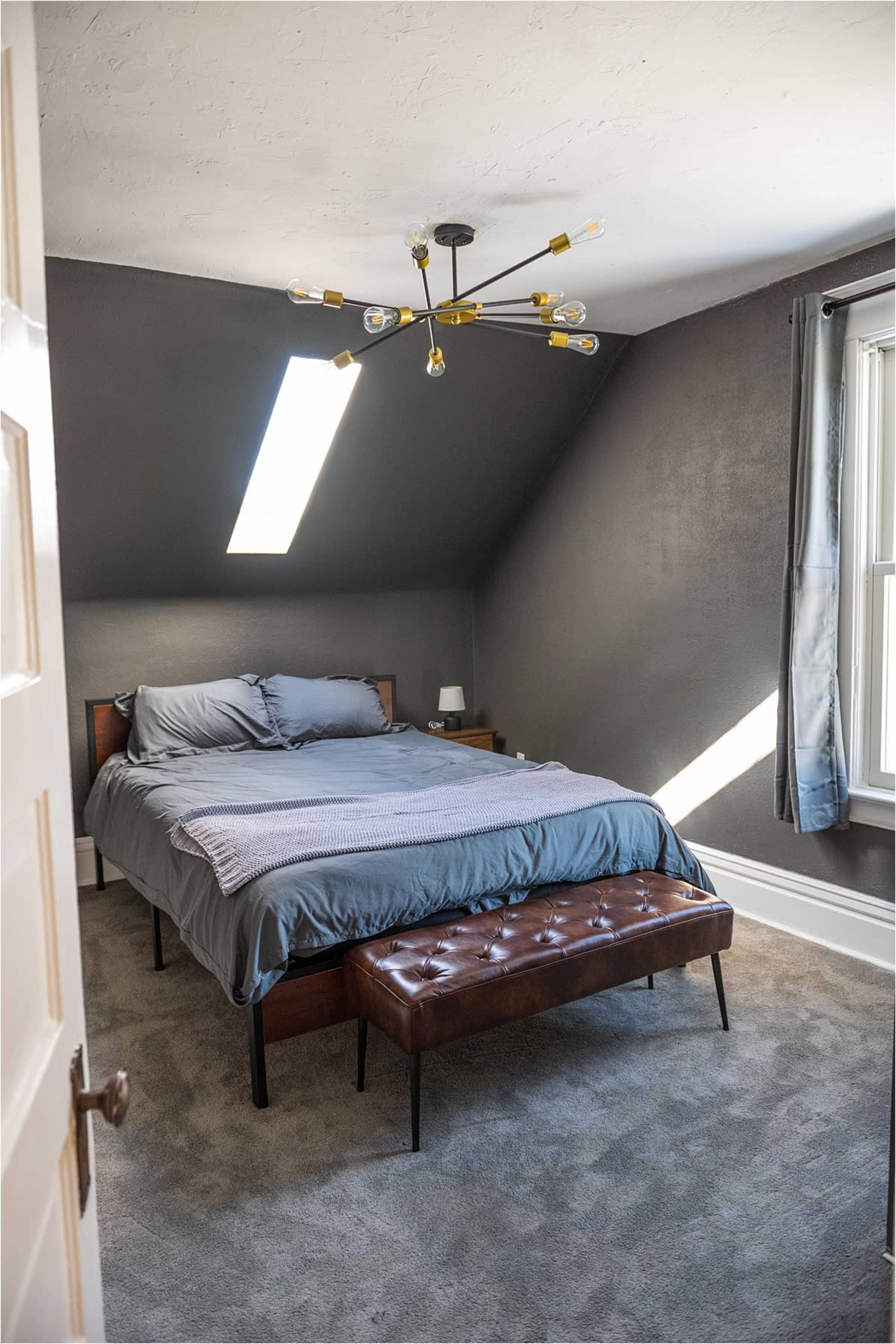 A minimalistic bedroom featuring a bed with gray bedding, a chandelier with exposed bulbs, and a tufted bench at the foot of the bed.