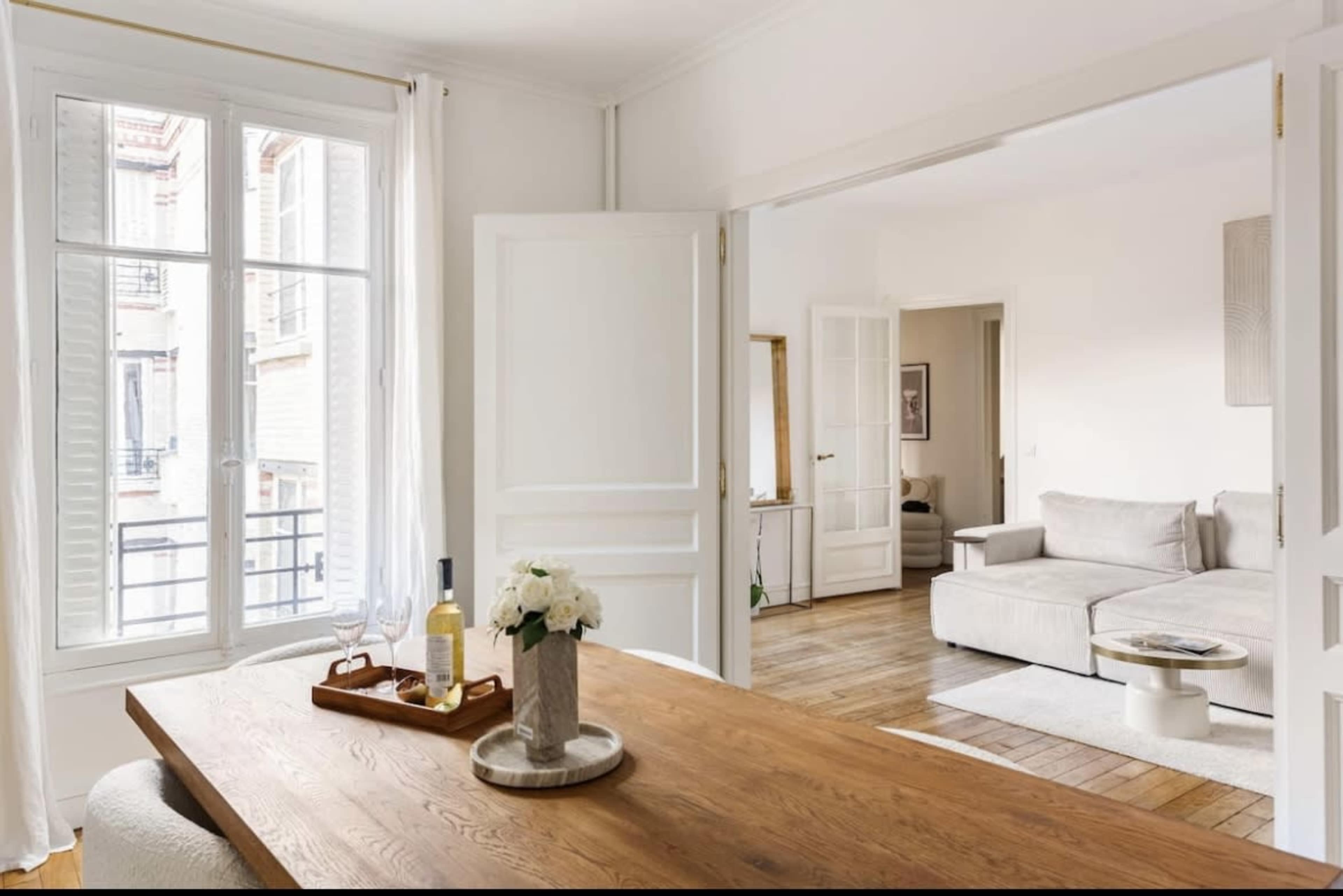 A bright, airy apartment with wooden floors, featuring a dining table with a vase of flowers and a view into a connected living room.