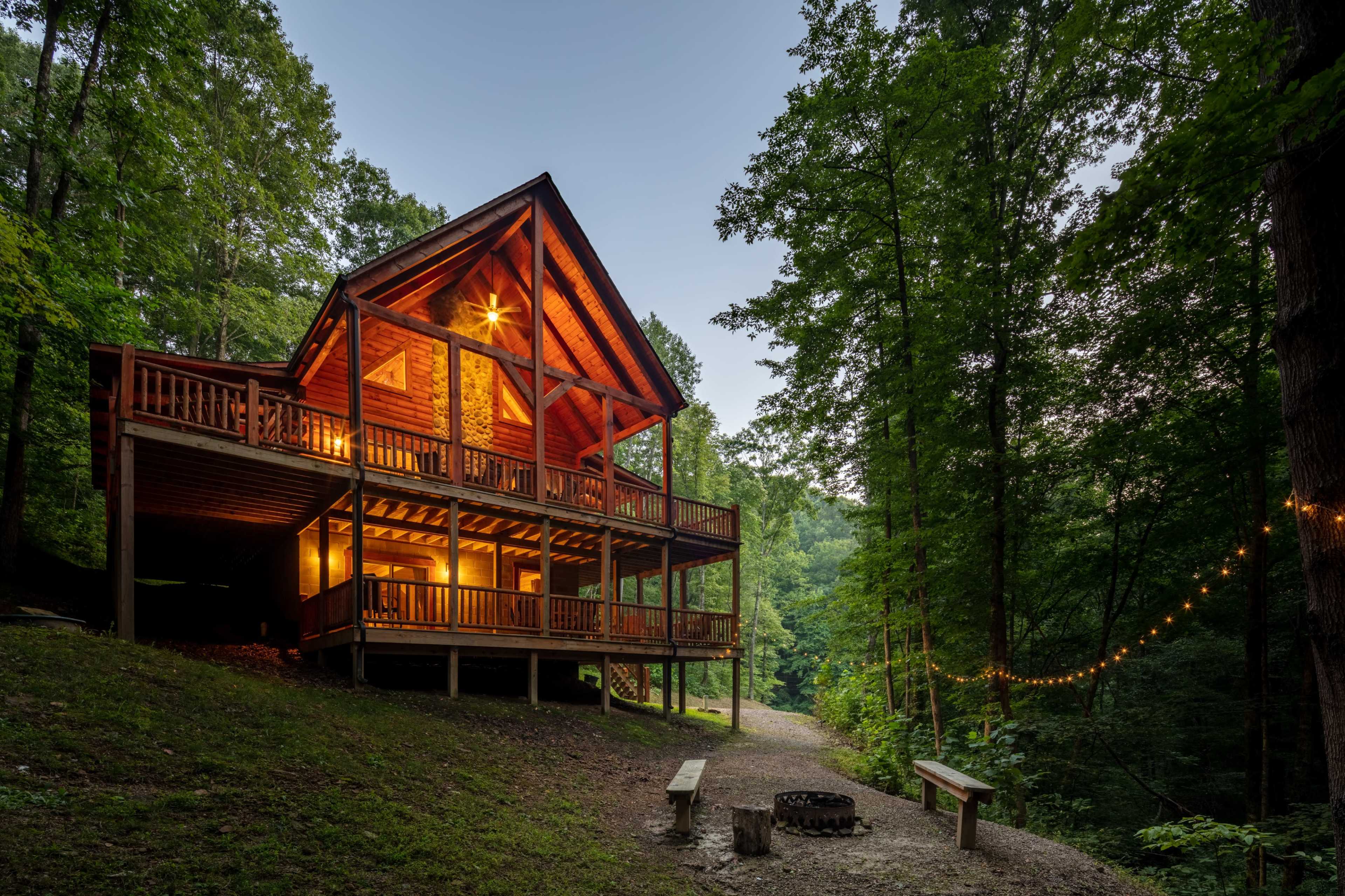 A large cabin with a wooden structure and balcony is surrounded by trees, with a pathway leading to a fire pit area.