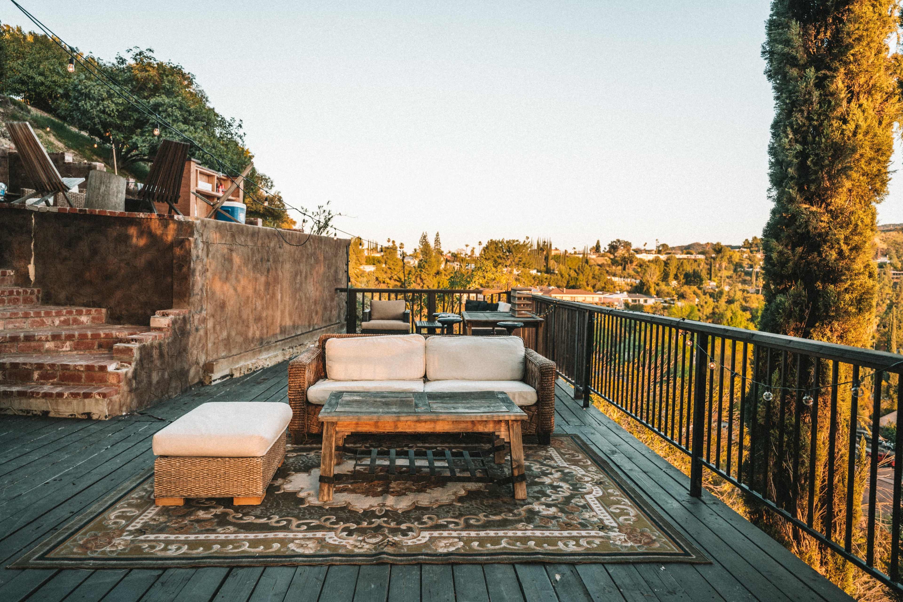 A wooden deck features a wicker sofa and ottoman on a patterned rug, overlooking a green landscape.