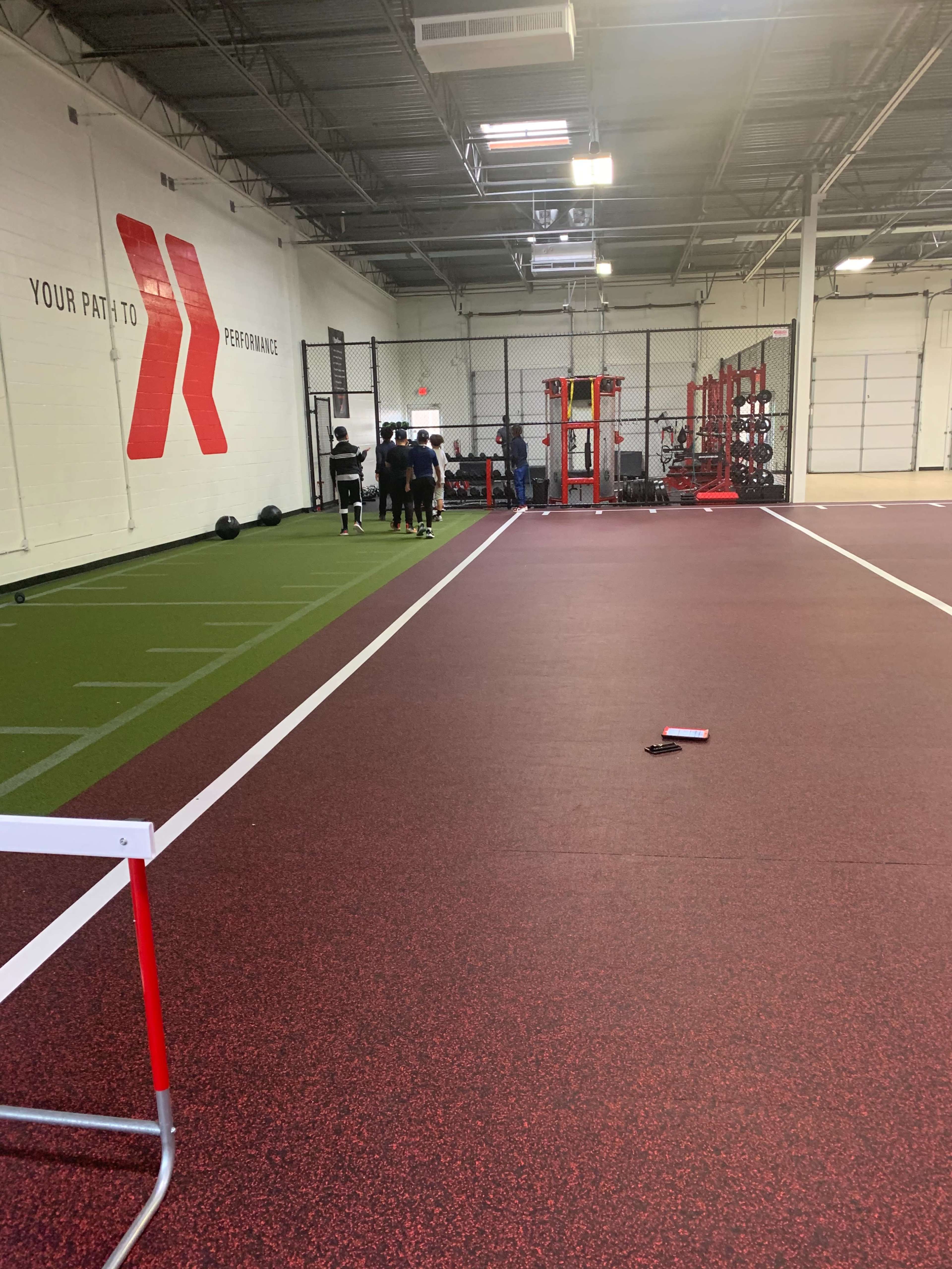 The image shows an indoor training facility with a red and black rubber floor, turf, exercise equipment, and a group of athletes lined up near the wall.