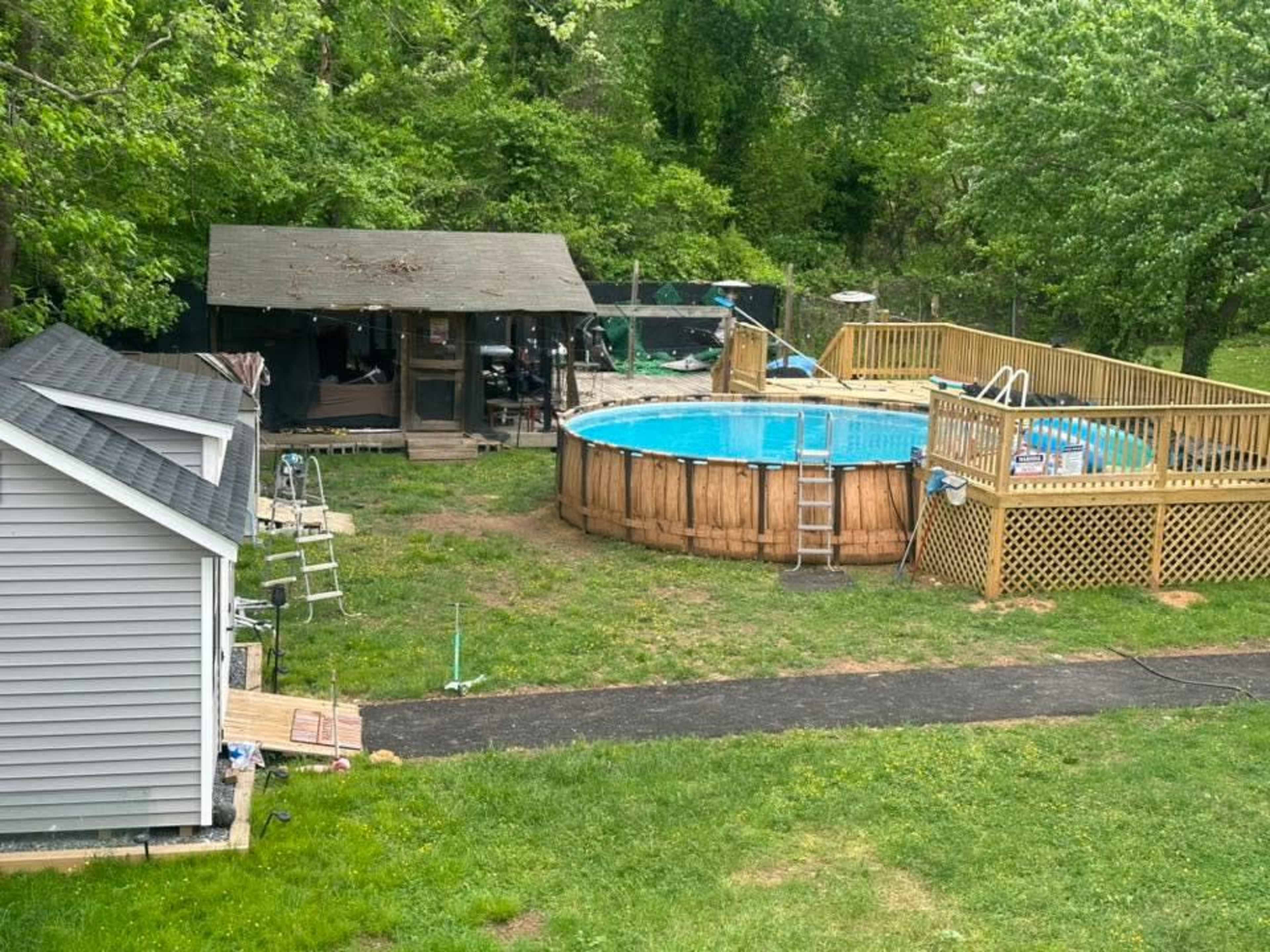 The image shows a backyard with a circular above-ground pool surrounded by a wooden deck, a shed, and a pathway leading through the grass.