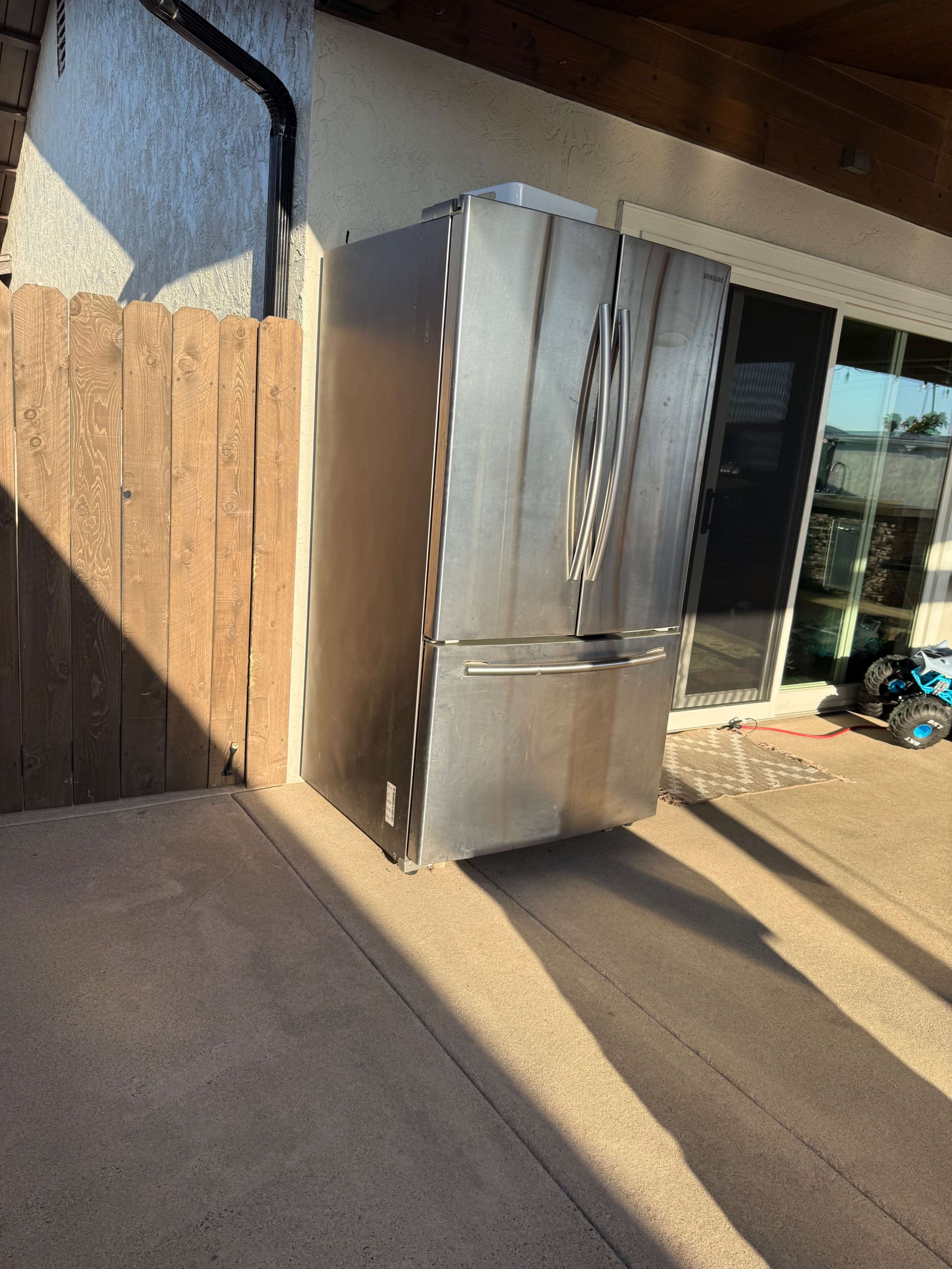 A stainless steel refrigerator stands on a patio next to a wooden fence and sliding glass doors.