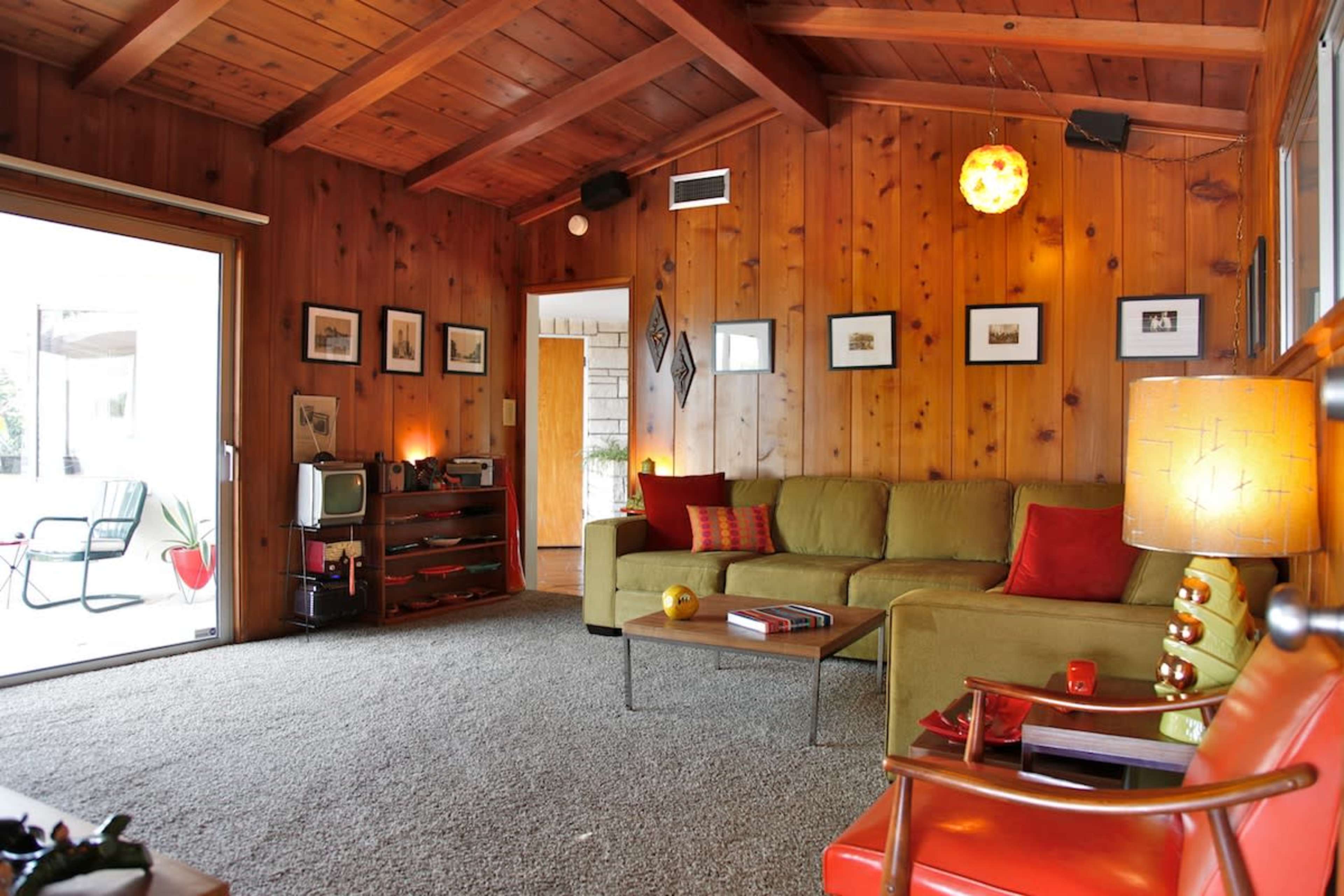 The image shows a cozy living room with wooden walls, a green sectional sofa, a coffee table, and a mix of framed photographs on the walls.