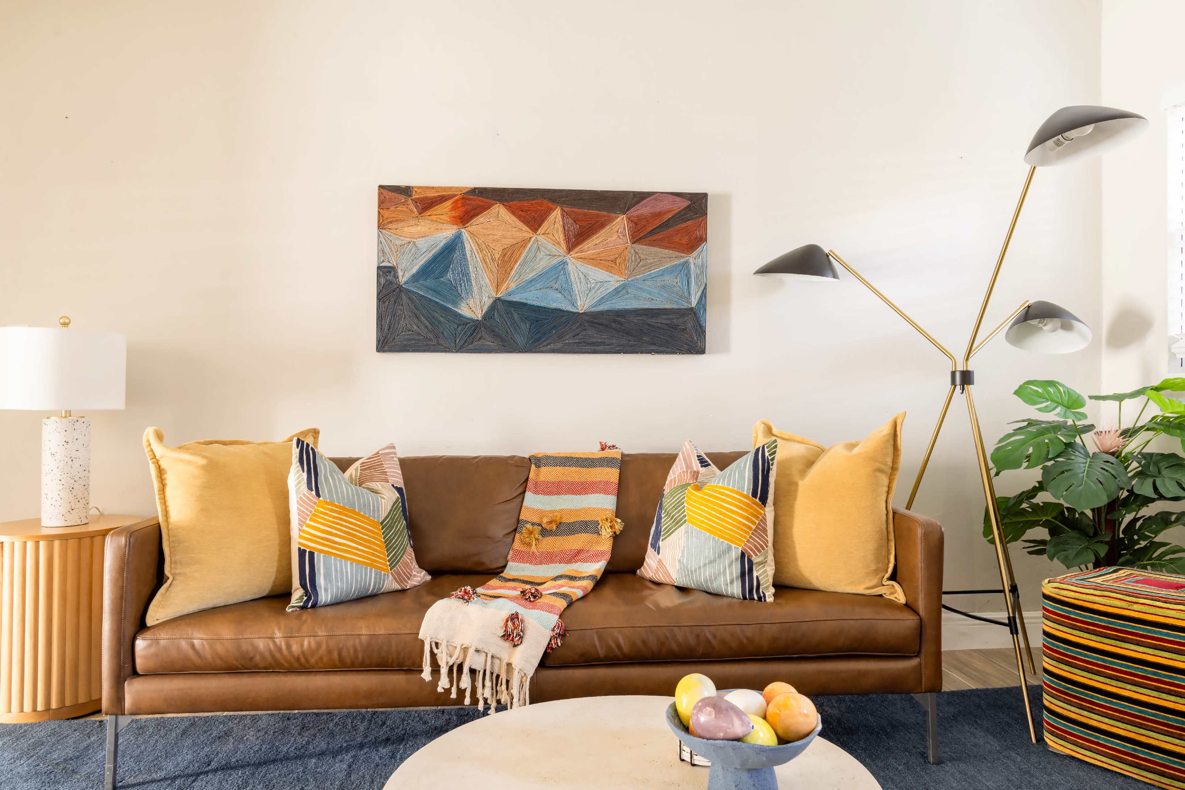 A brown leather sofa with colorful patterned pillows is placed against a white wall, accompanied by a round coffee table and a tall lamp, with a large abstract painting above and a green plant nearby.