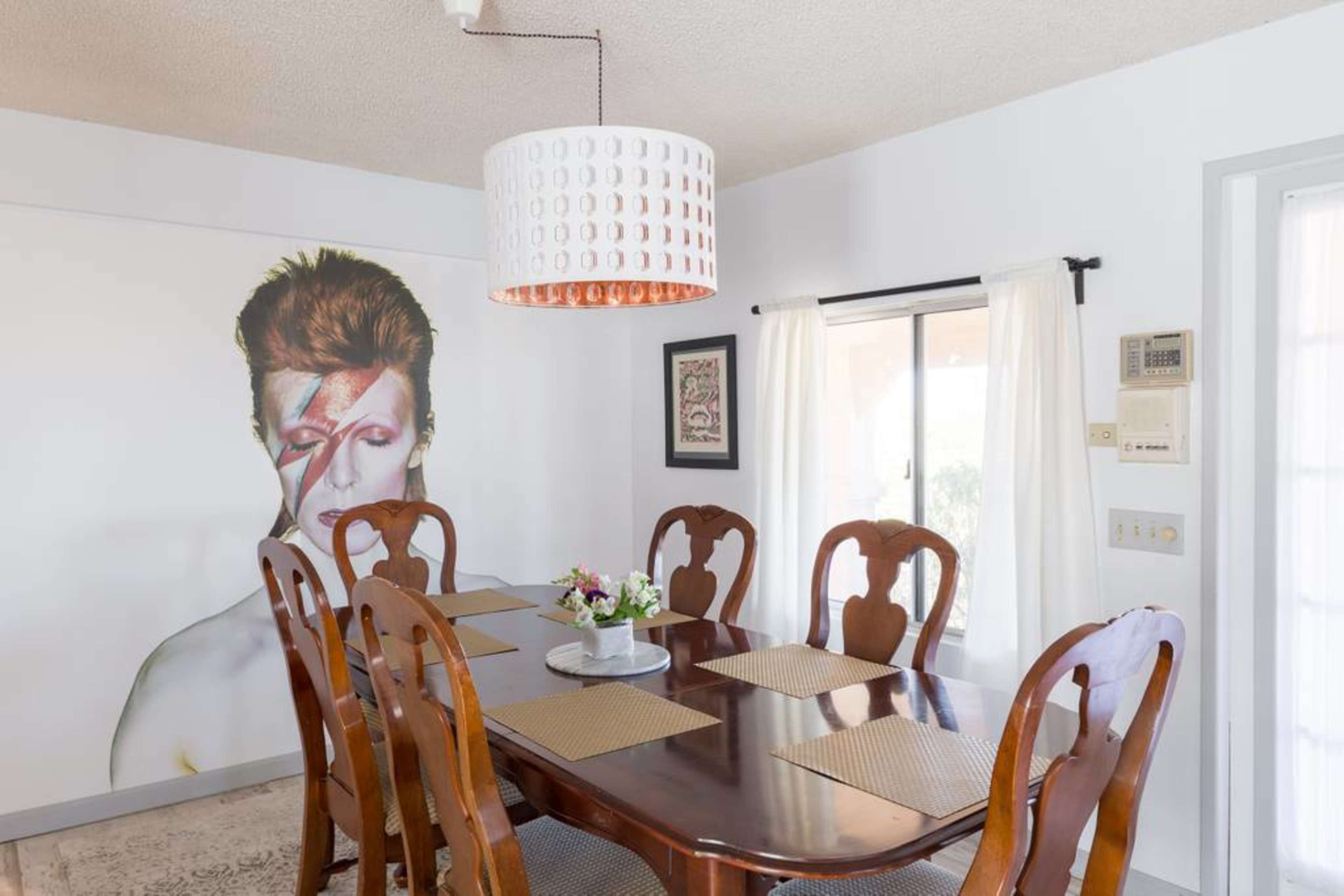 A dining room with a large wooden table surrounded by chairs, featuring a mural of a person with a distinctive makeup design on the wall.
