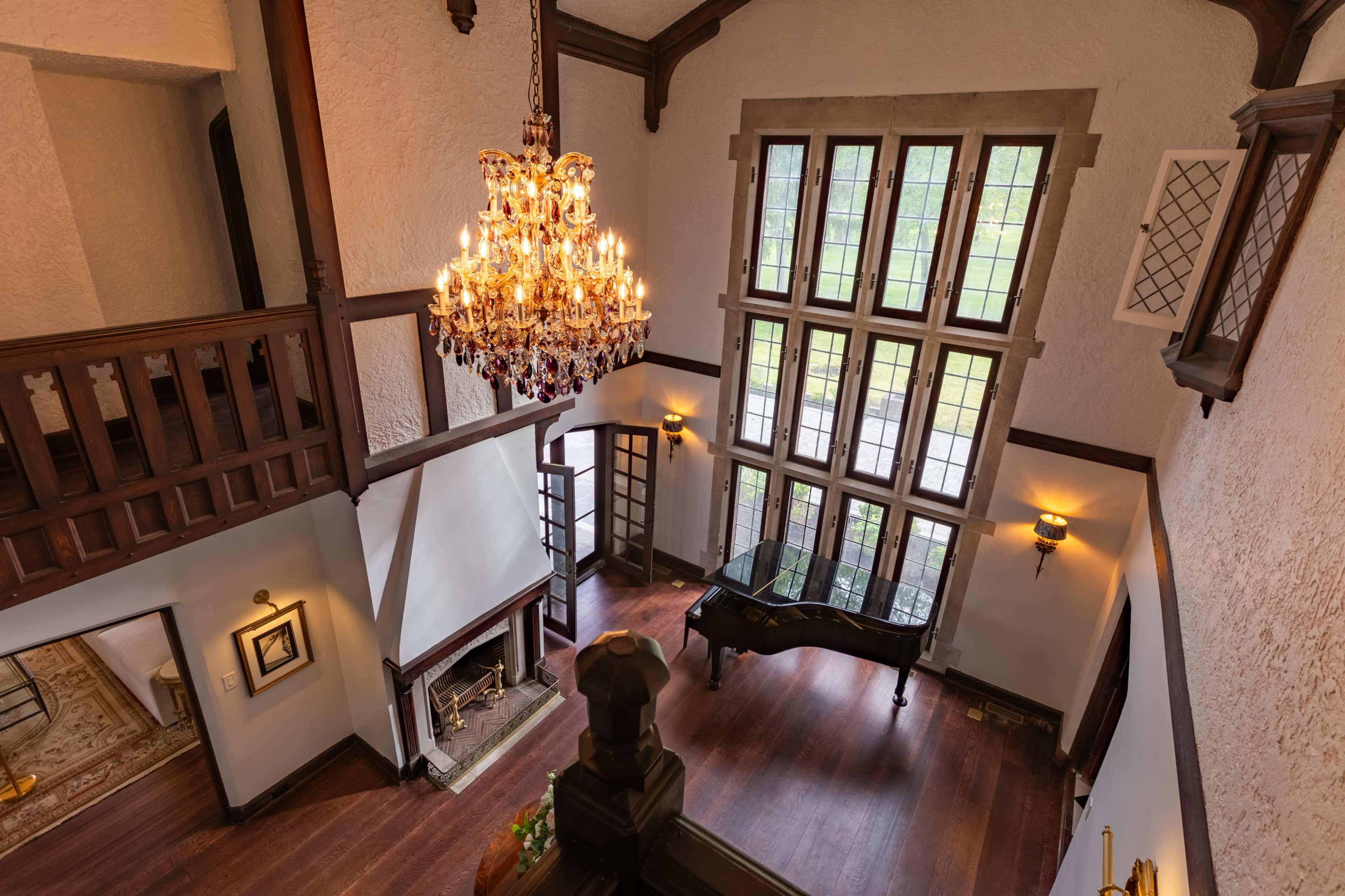 The image shows a spacious interior with a high ceiling, featuring a large chandelier, a grand piano near the windows, and a fireplace.