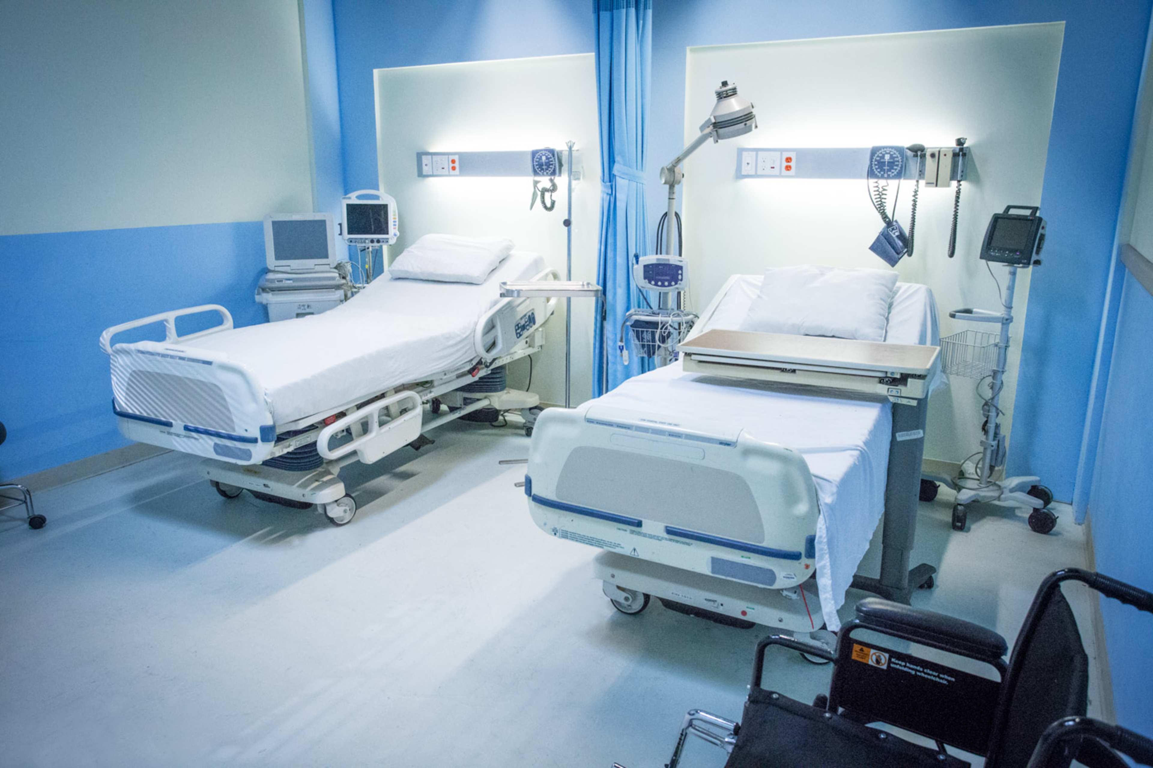 A hospital room with two empty beds, medical equipment, and a wheelchair in the corner.