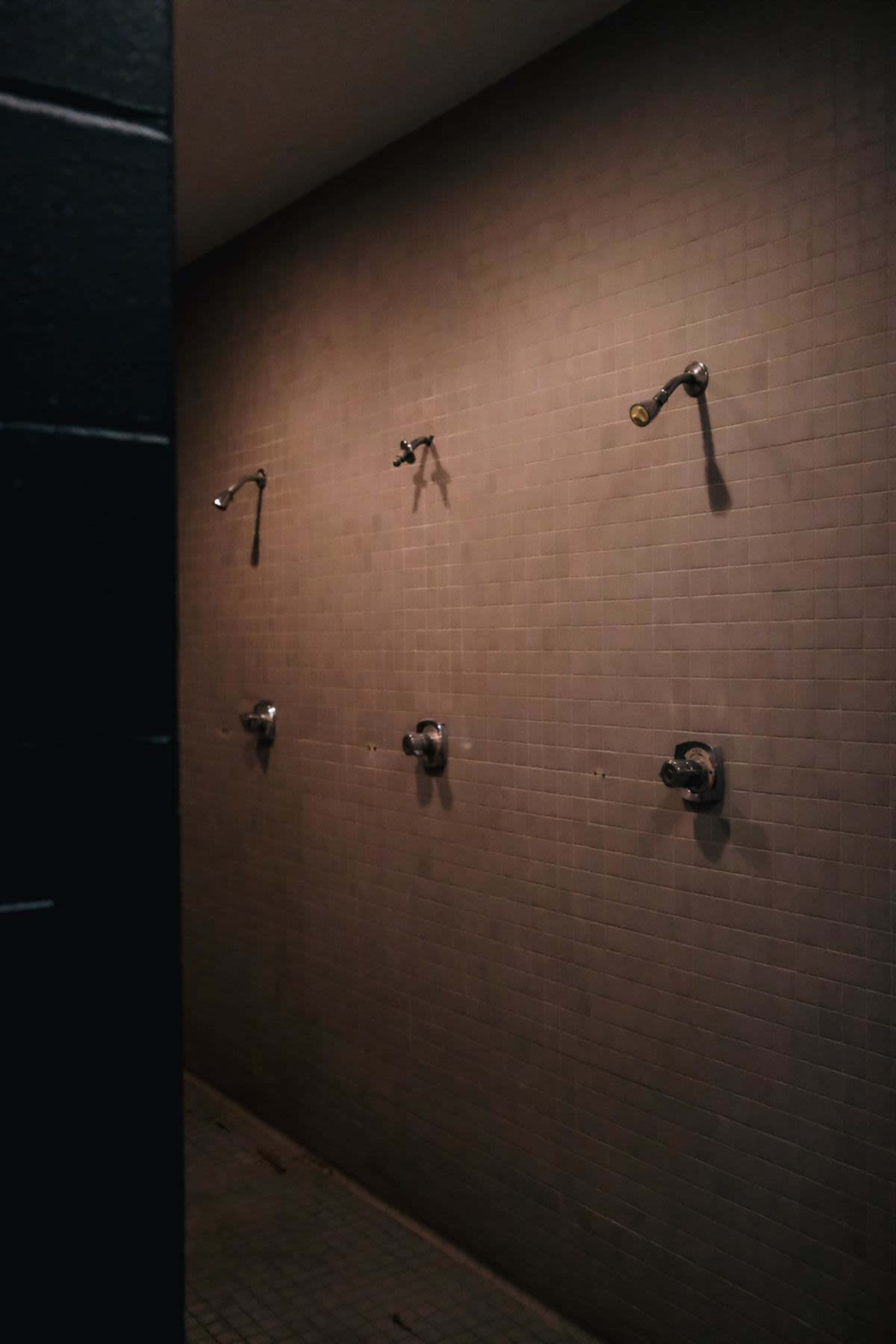 The image shows a wall with three showerheads mounted above a tiled floor in a dimly lit space.