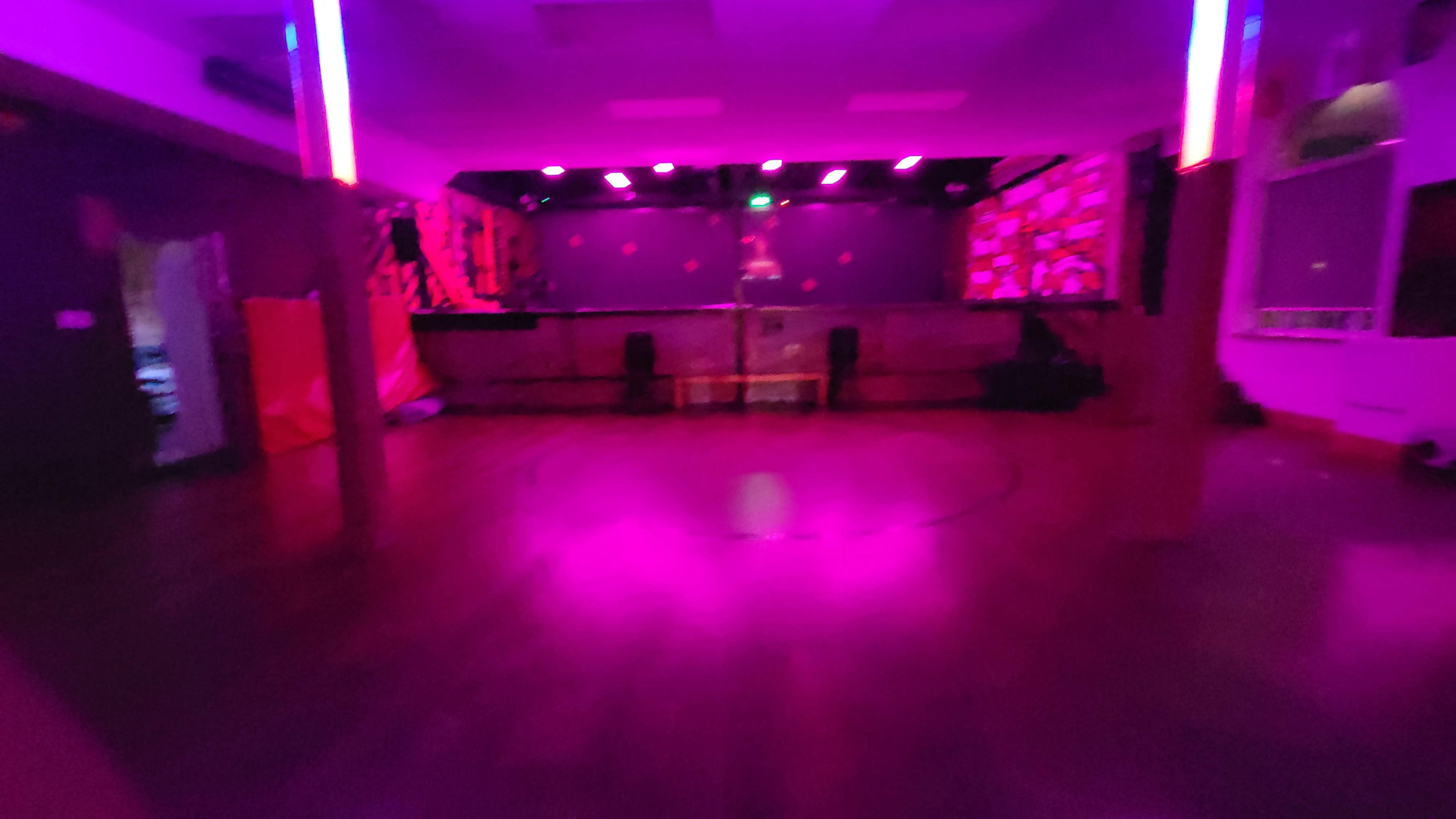 The image depicts a dimly lit room with purple lighting, showcasing an empty dance floor surrounded by walls featuring colorful decor.