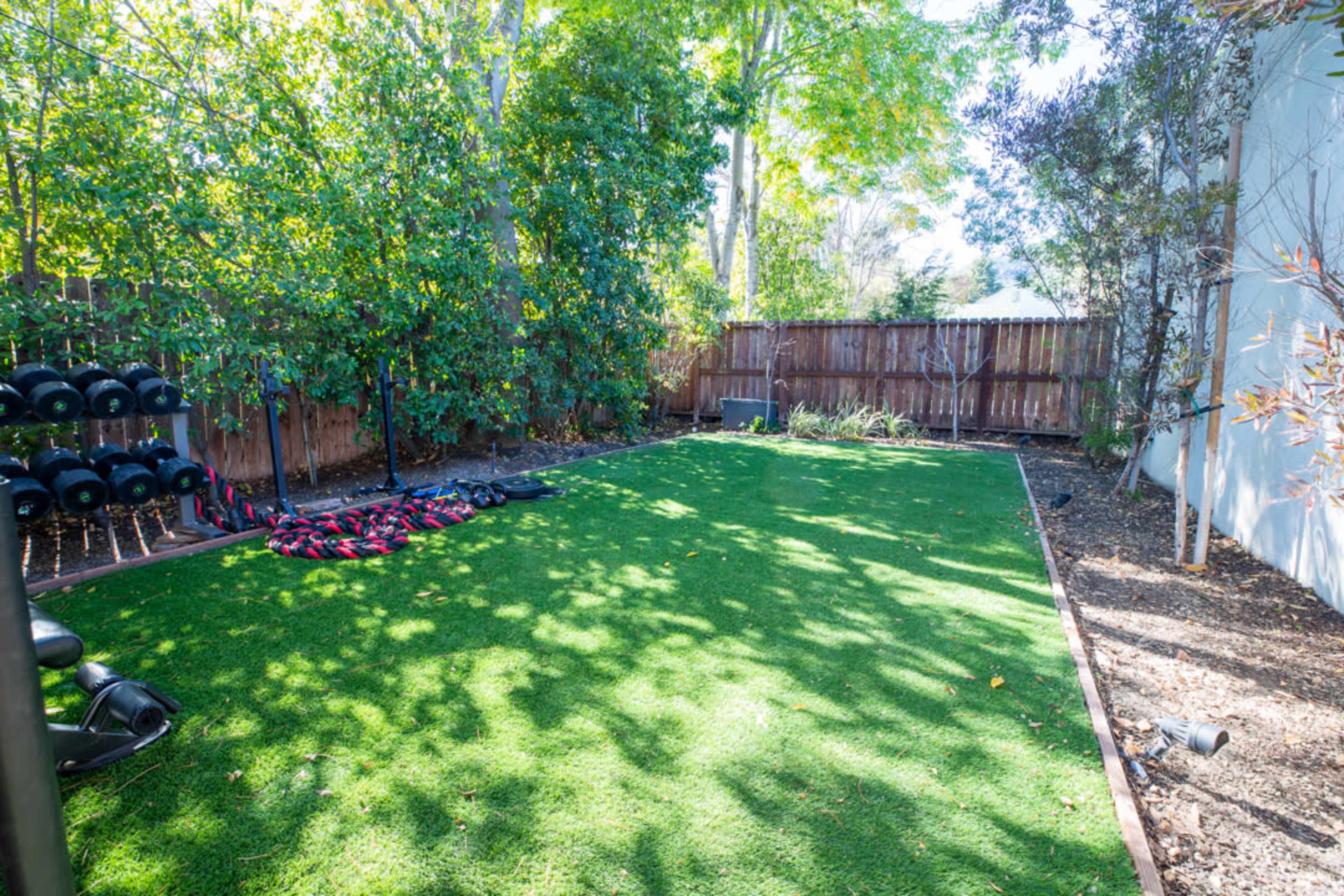 A fitness area with artificial grass, dumbbells arranged on the left, and a fenced garden space with shrubbery in the background.
