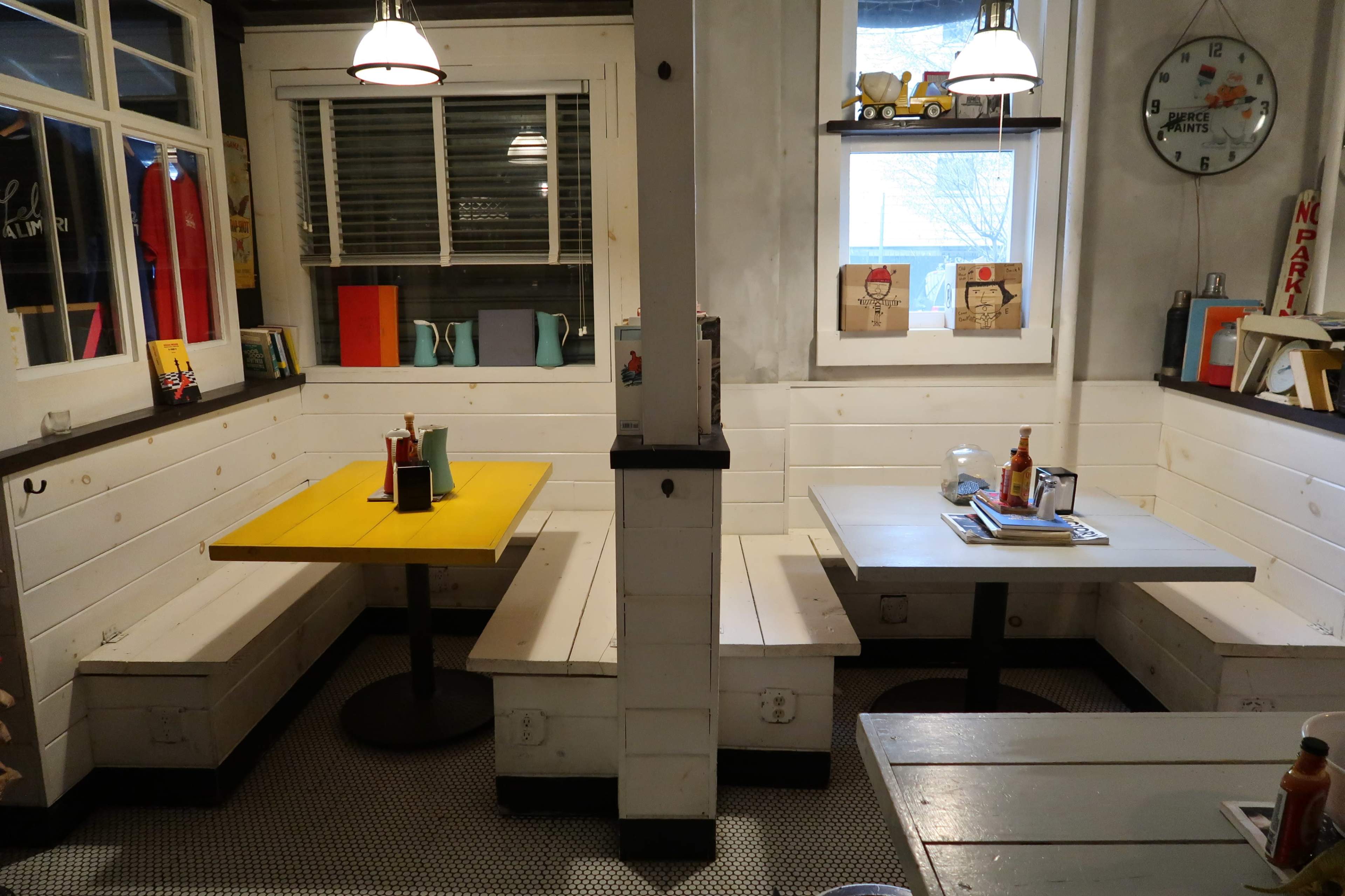 A restaurant interior with two tables, one yellow and one gray, surrounded by white wooden benches and decorated with various items on the walls.