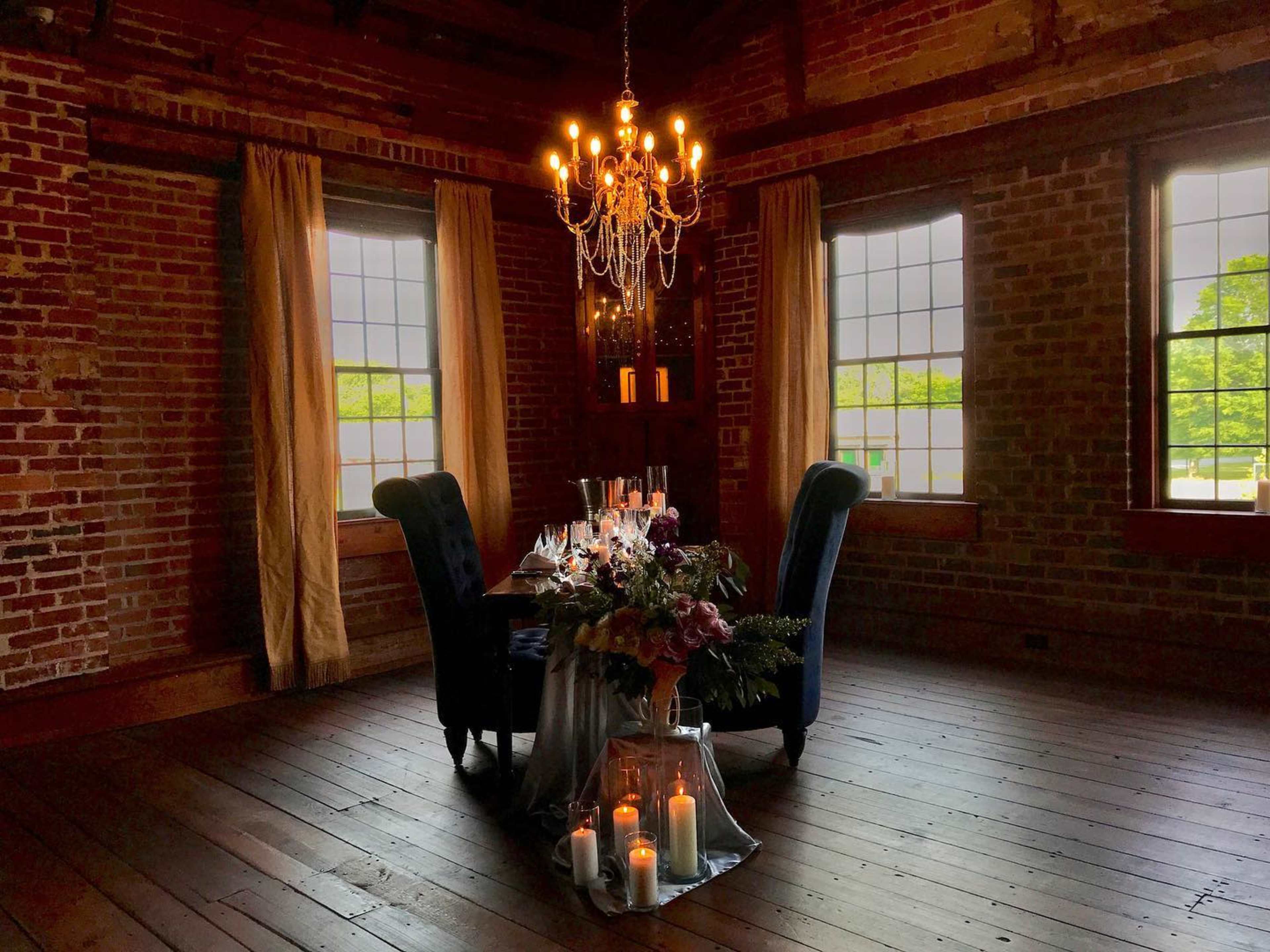 A dining table set for two is illuminated by a chandelier in a room with brick walls and large windows.