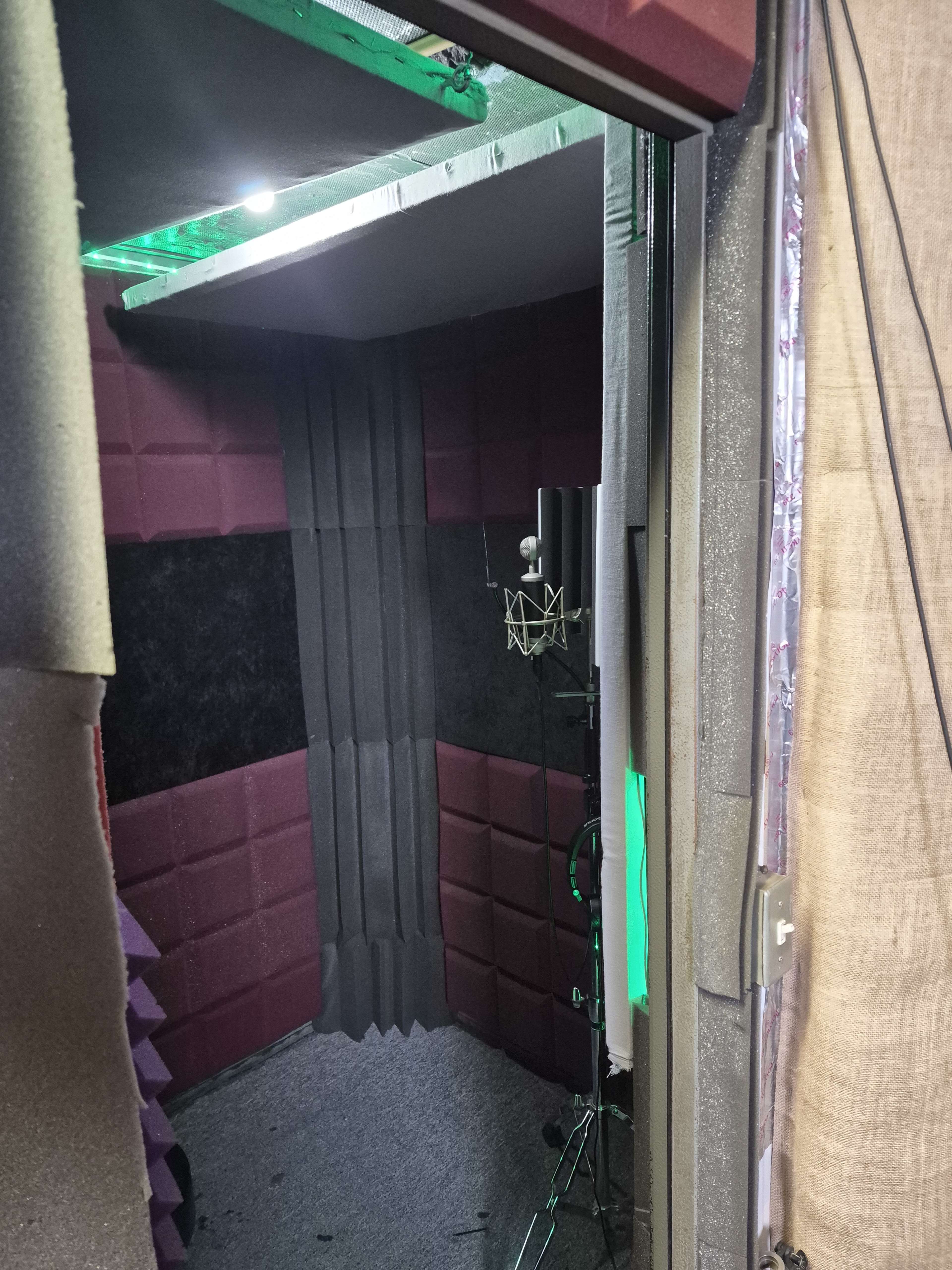 The image shows the interior of a soundproof recording booth featuring acoustic panels and a microphone setup.