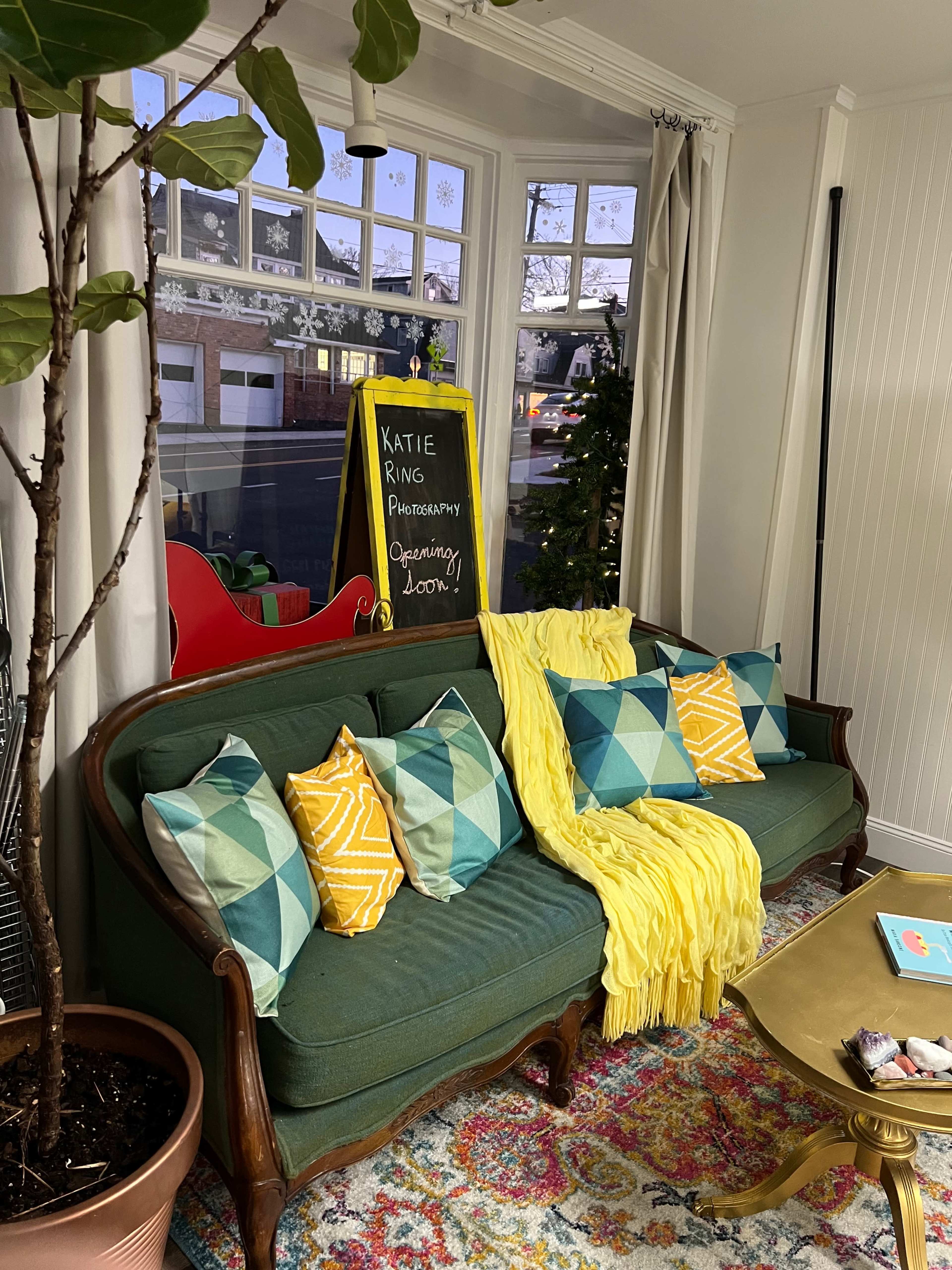 A green sofa with patterned cushions and a yellow throw is positioned in a brightly lit room near a chalkboard sign announcing a photography opening, with decorative plants and a colorful rug visible.