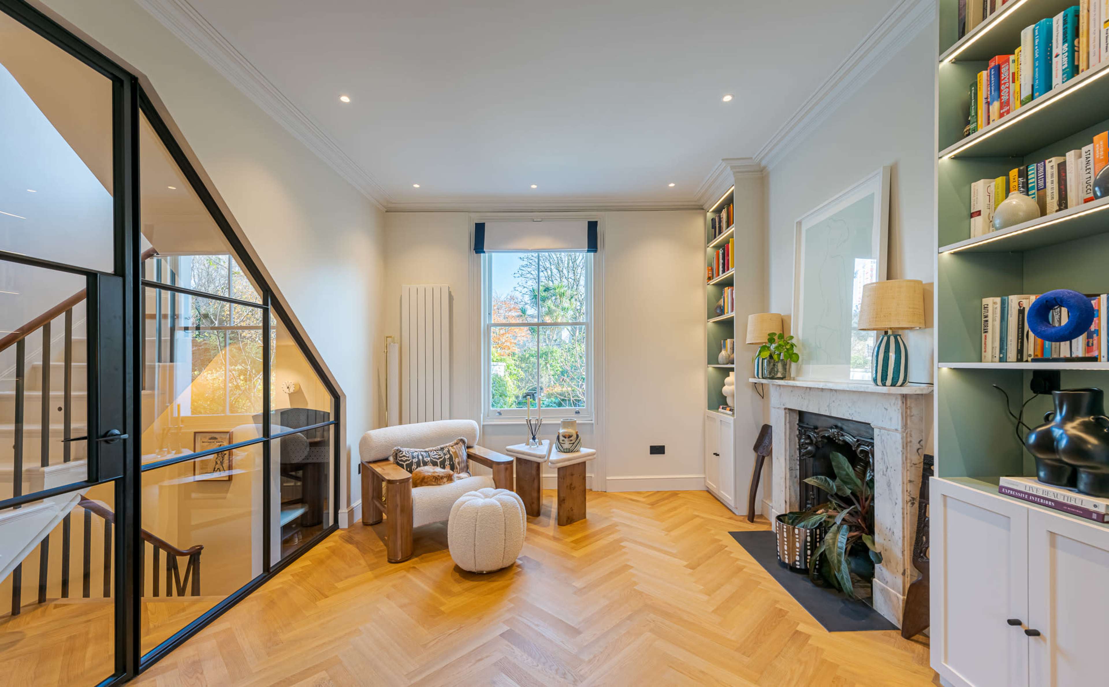 The image shows a modern living room with a cozy seating area, a bookshelf, and large windows allowing natural light to brighten the space.