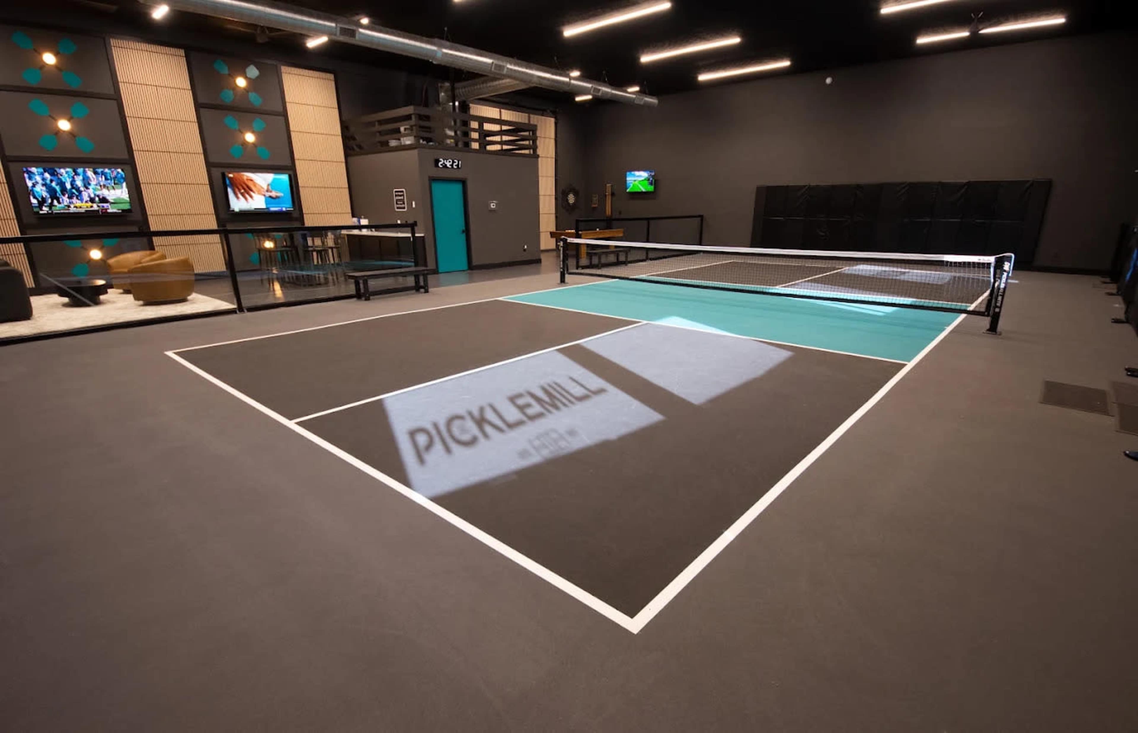 The image shows an indoor sports facility with two pickleball courts, equipped with a net and marked lines, and features a modern design with TVs and seating areas visible in the background.