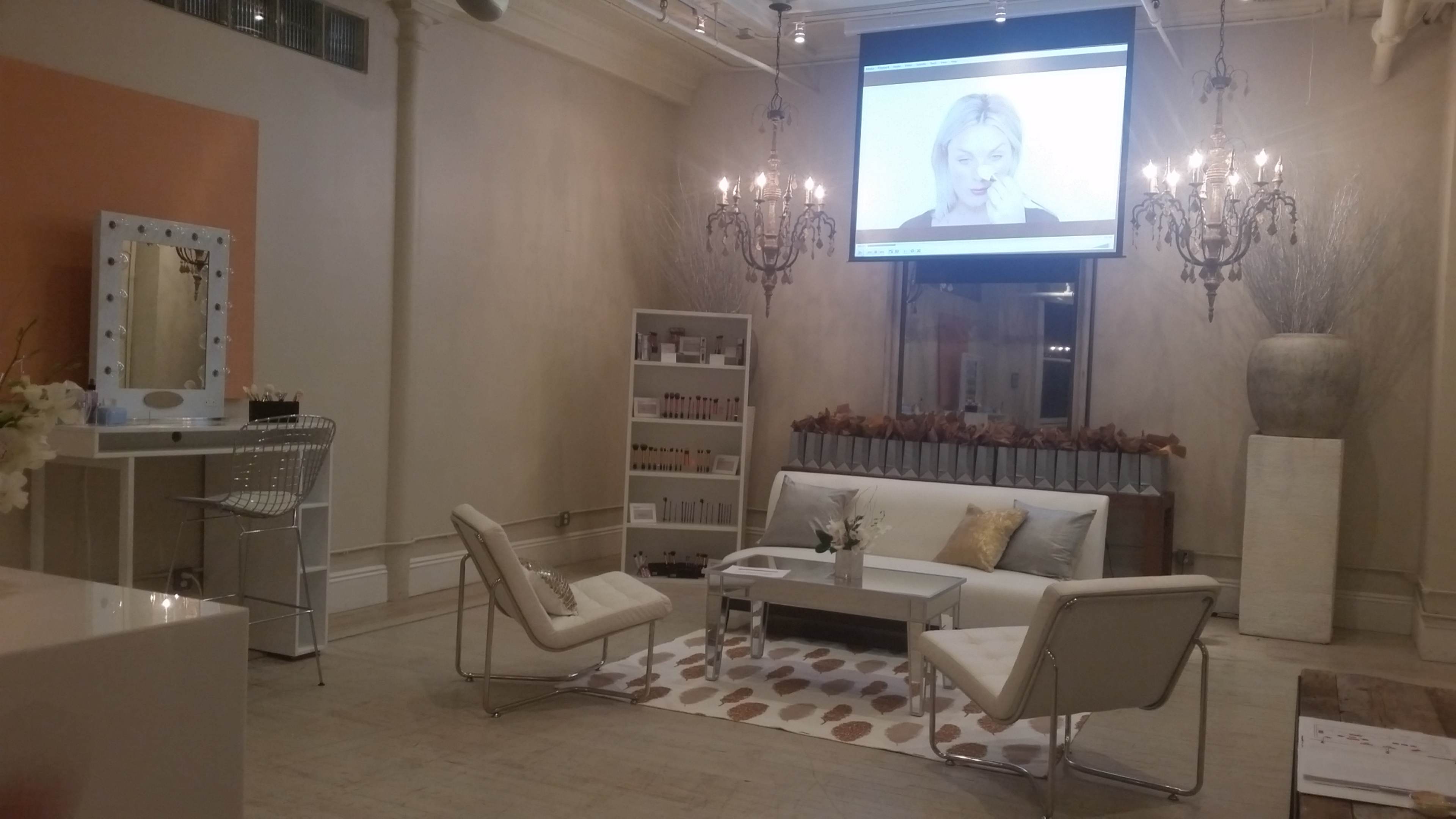 The image shows a brightly lit salon or spa interior featuring a seating area with modern chairs, a coffee table, and a large screen displaying a video.