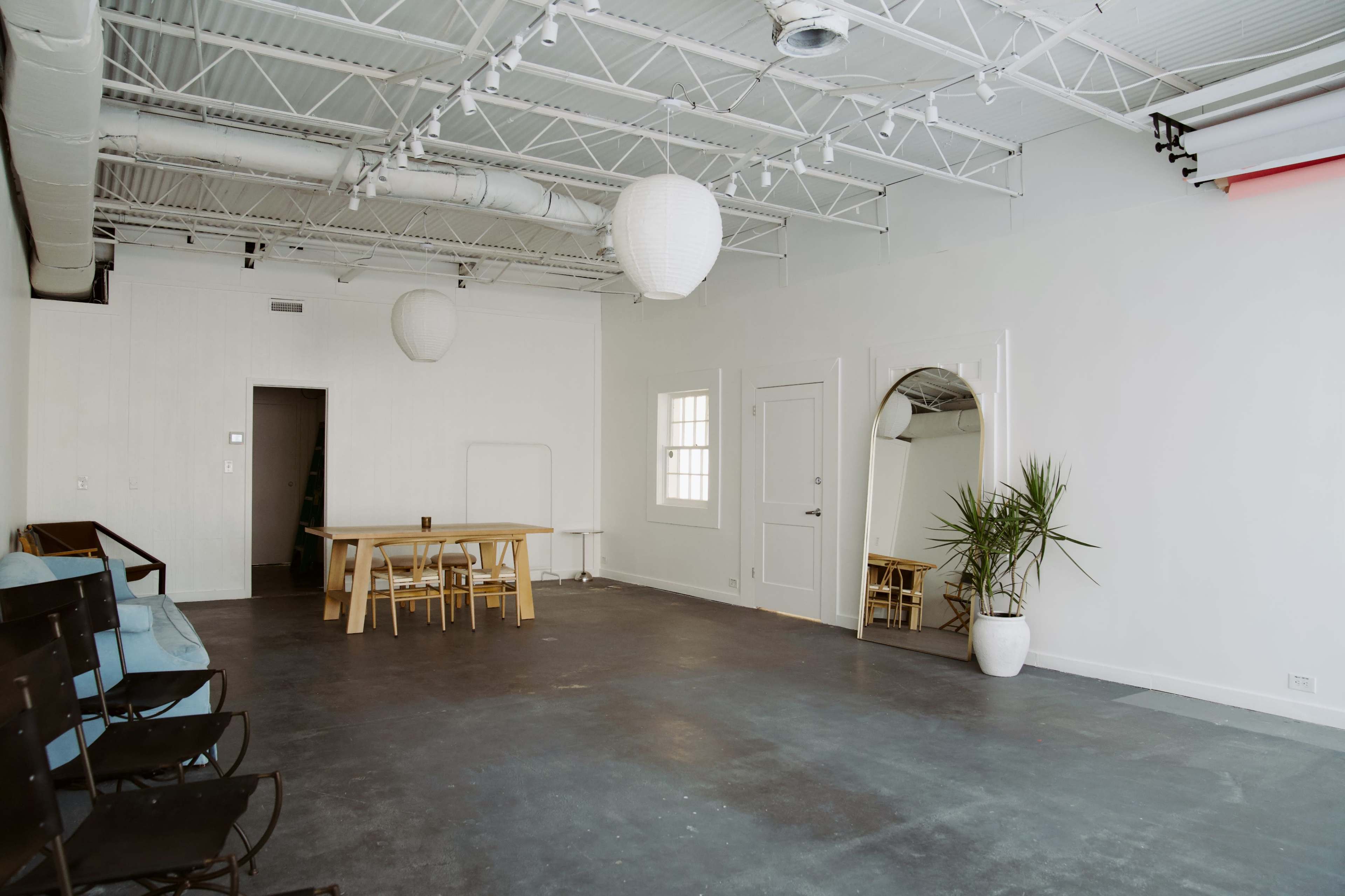 The image shows a spacious, minimally furnished room with a long wooden table, a few chairs, a potted plant, and large windows allowing natural light to enter.