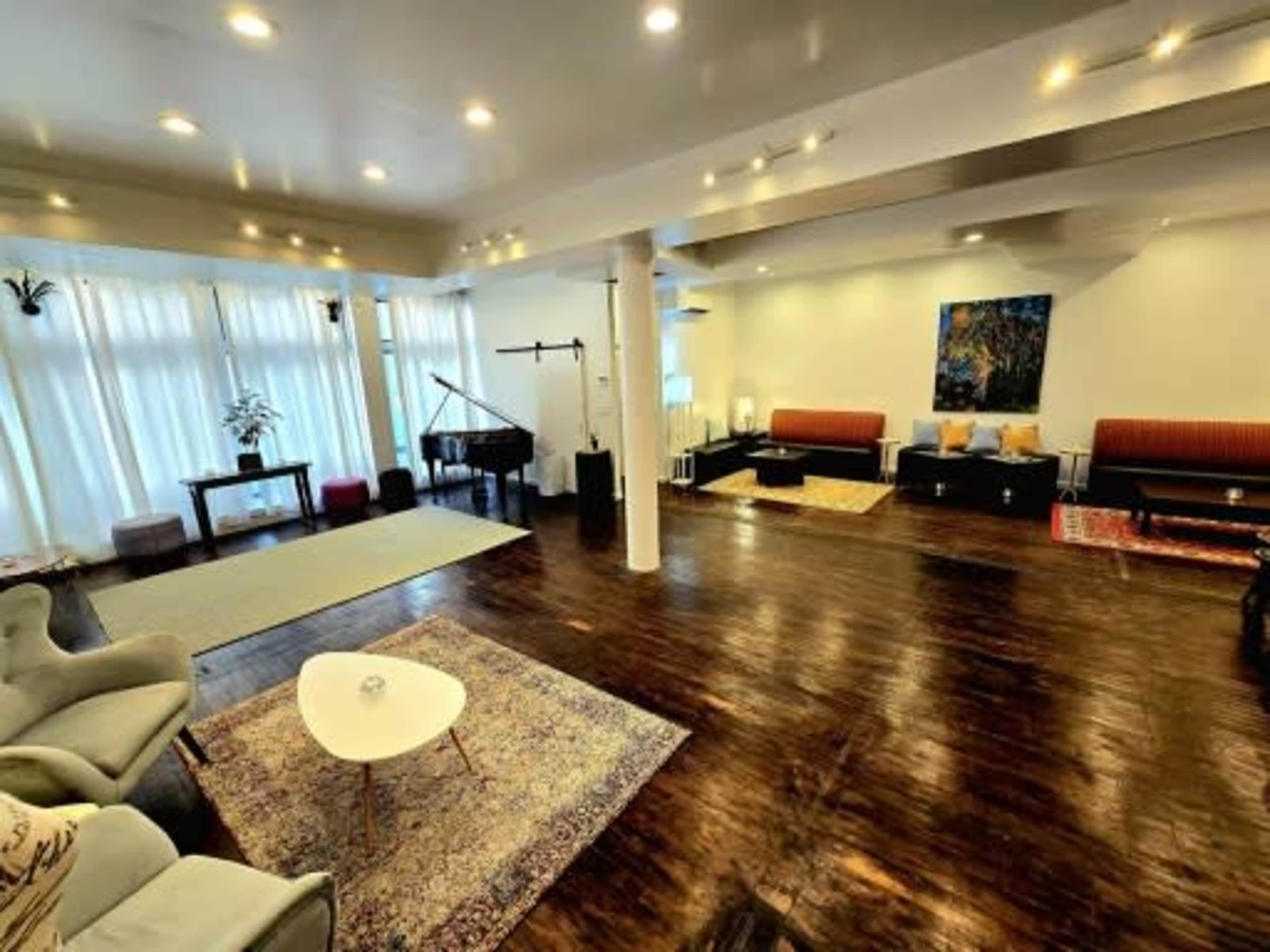 The image shows a spacious and well-lit living area featuring a grand piano, a light-colored area rug, comfortable seating arrangements, and large windows with sheer curtains.