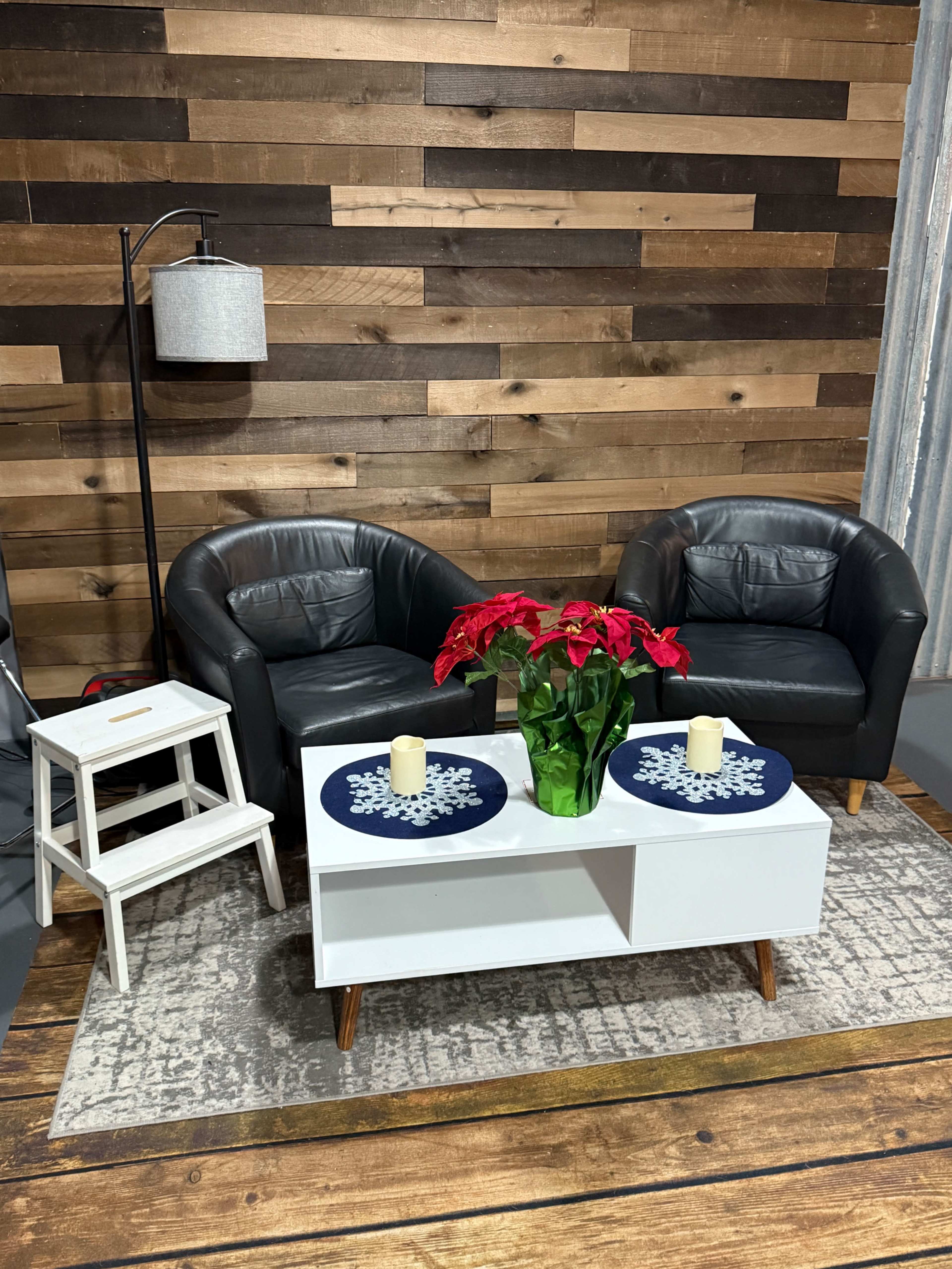 The image shows a cozy living room setup with two black leather chairs, a small white side table, a white coffee table decorated with blue snowflake mats and candles, and a planter with red poinsettias, all against a backdrop of wooden planks.