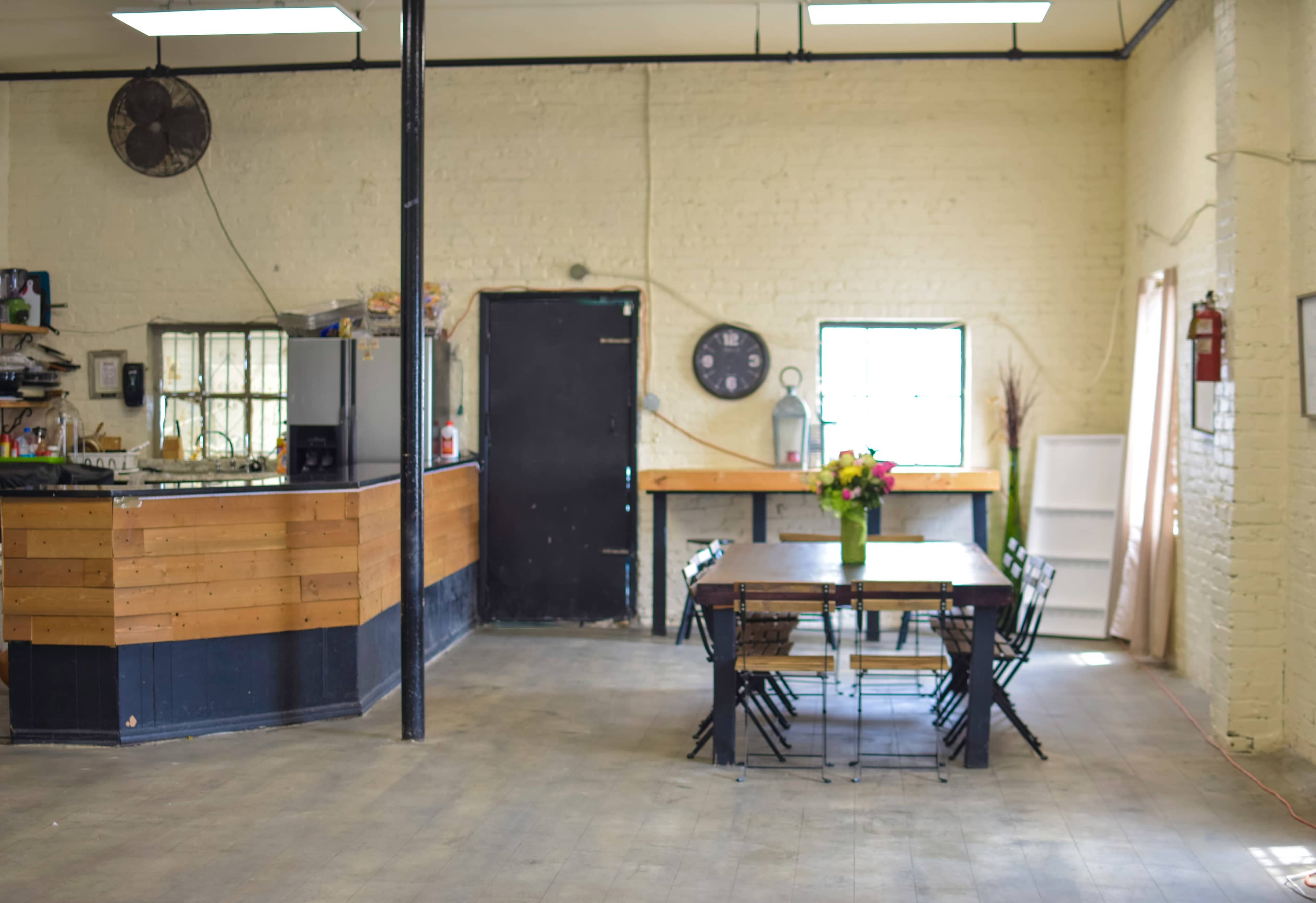 A spacious room with a dining area featuring a large table surrounded by chairs, a kitchenette to the left, and a doorway leading to another space.