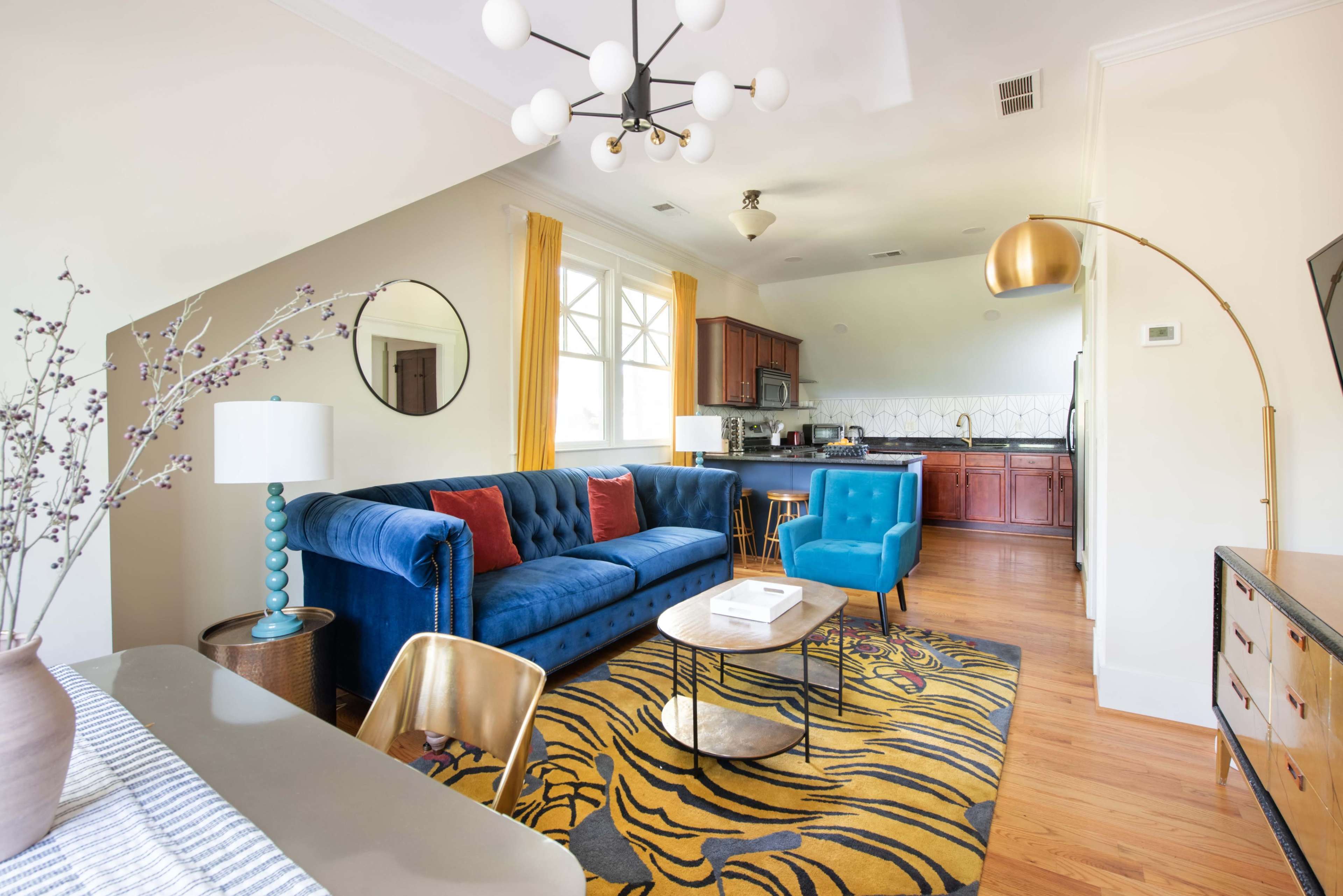 The image shows a modern living room with a blue velvet sofa, two teal chairs, a circular dining table, and a kitchen area in the background.