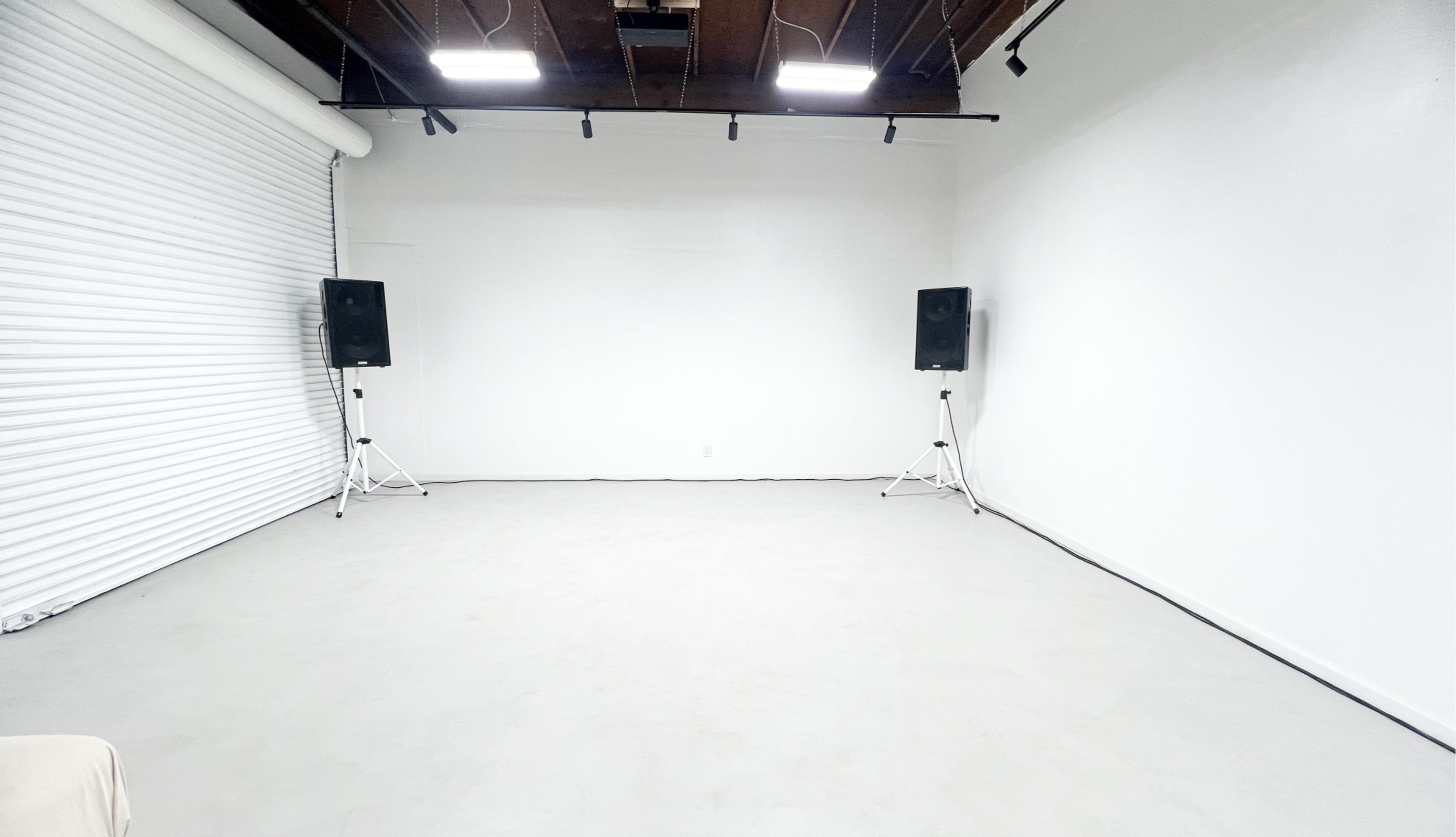 An empty room with white walls and a concrete floor, featuring two speakers mounted on stands against one wall.