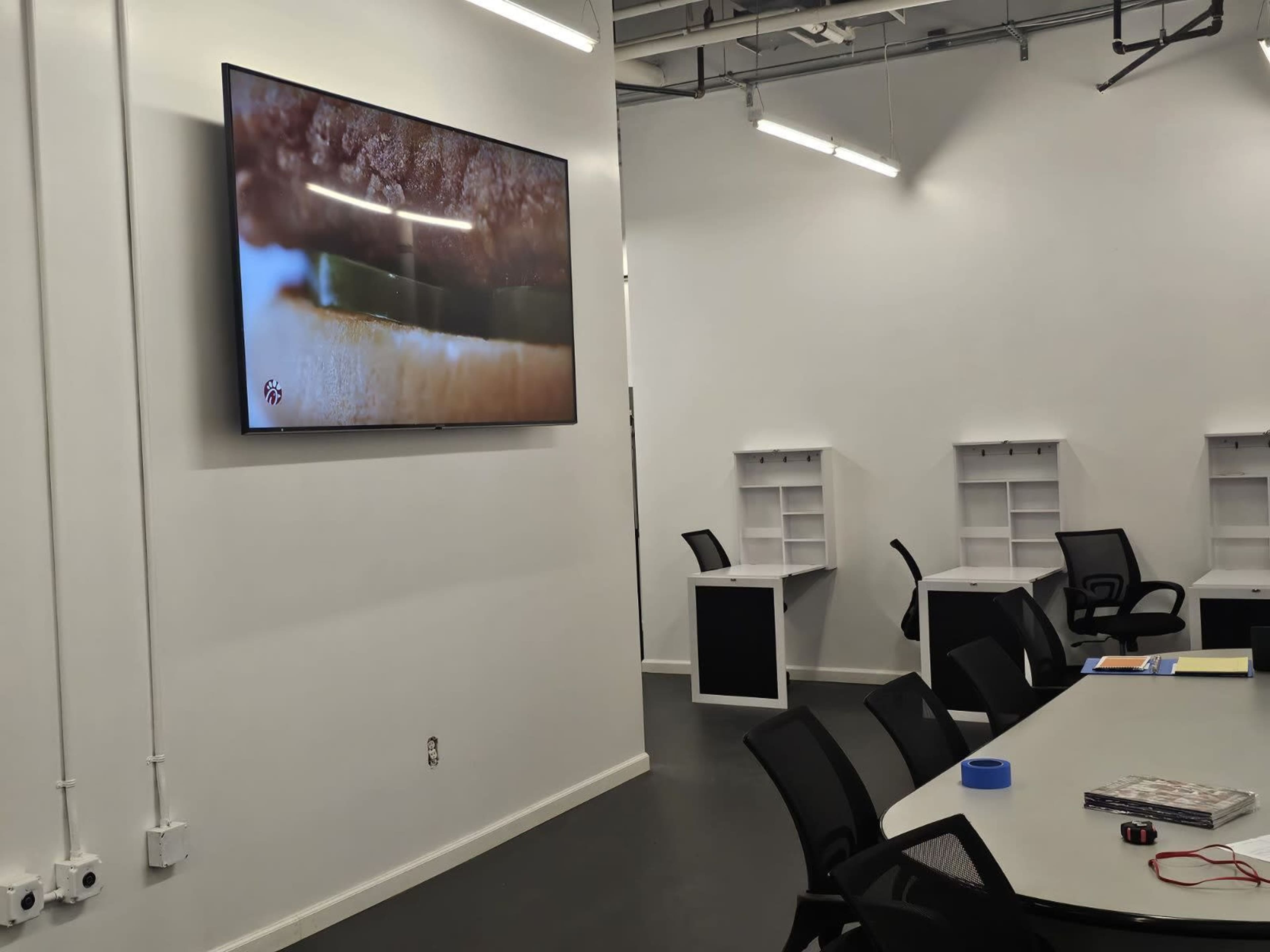 A large screen displays a close-up image in a modern, sparsely furnished meeting room with a long table and black chairs.