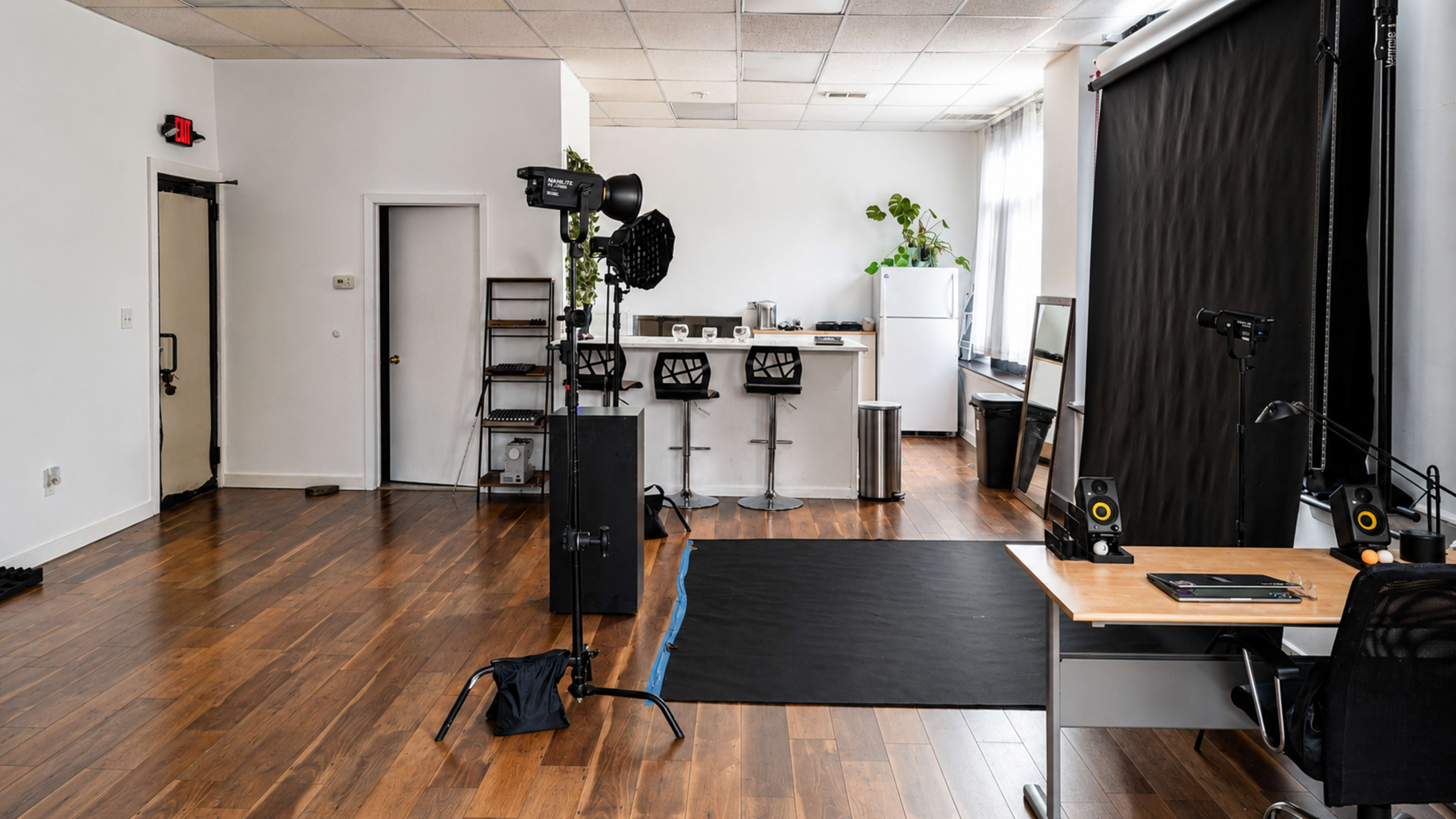 The image shows a spacious studio with wooden flooring that includes a photography setup, a workspace with a desk and chair, and a kitchenette area in the background.