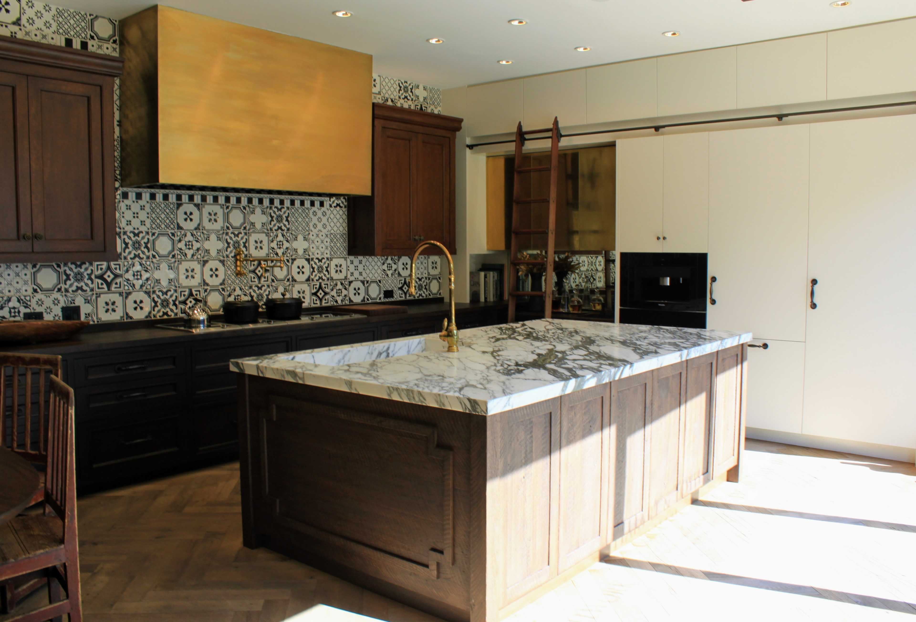A modern kitchen with a large marble island, dark wooden cabinets, patterned tile backsplash, and a gold hood above the stove.