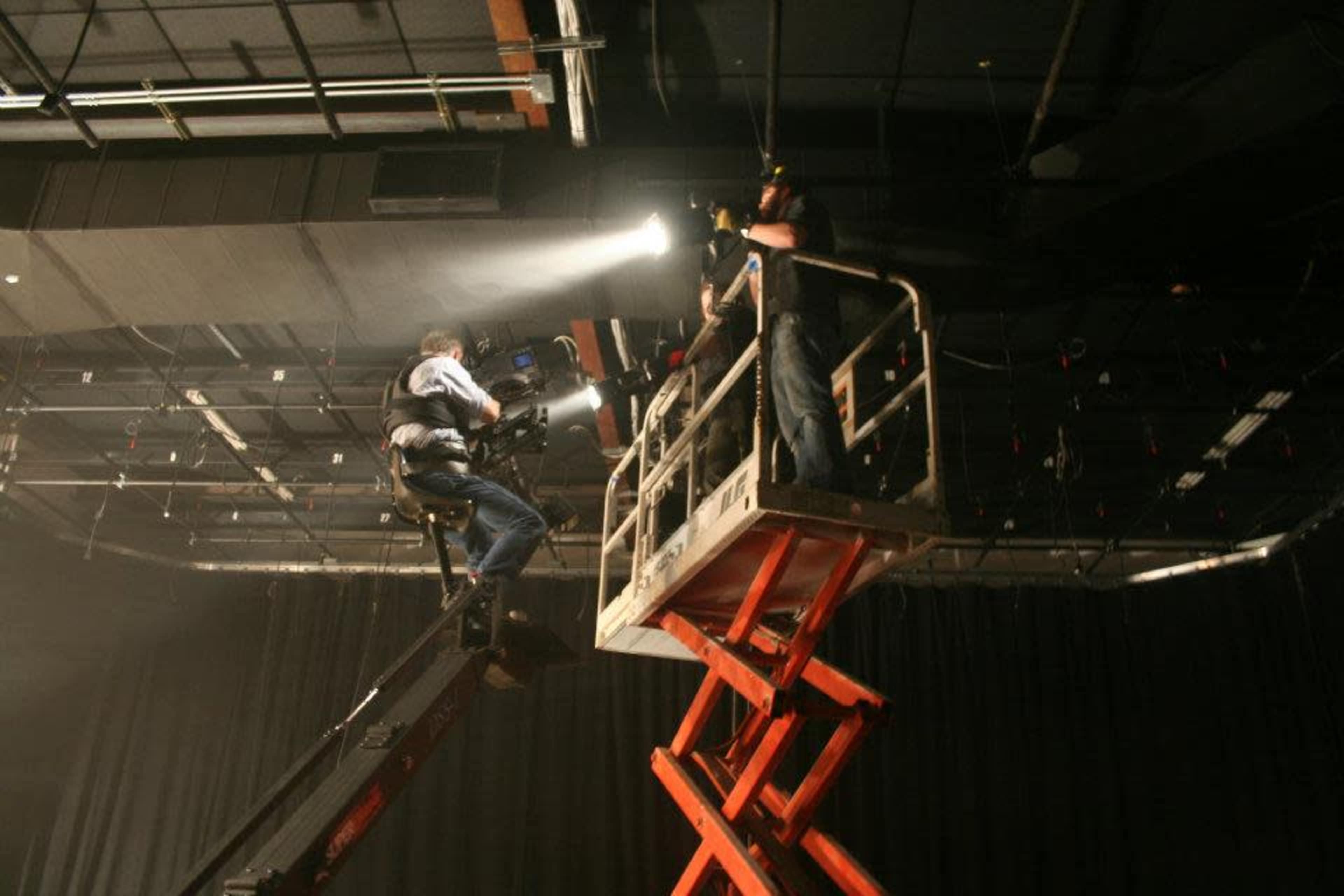 A crew member operates a camera from a raised platform while another person adjusts equipment in a dimly lit studio.