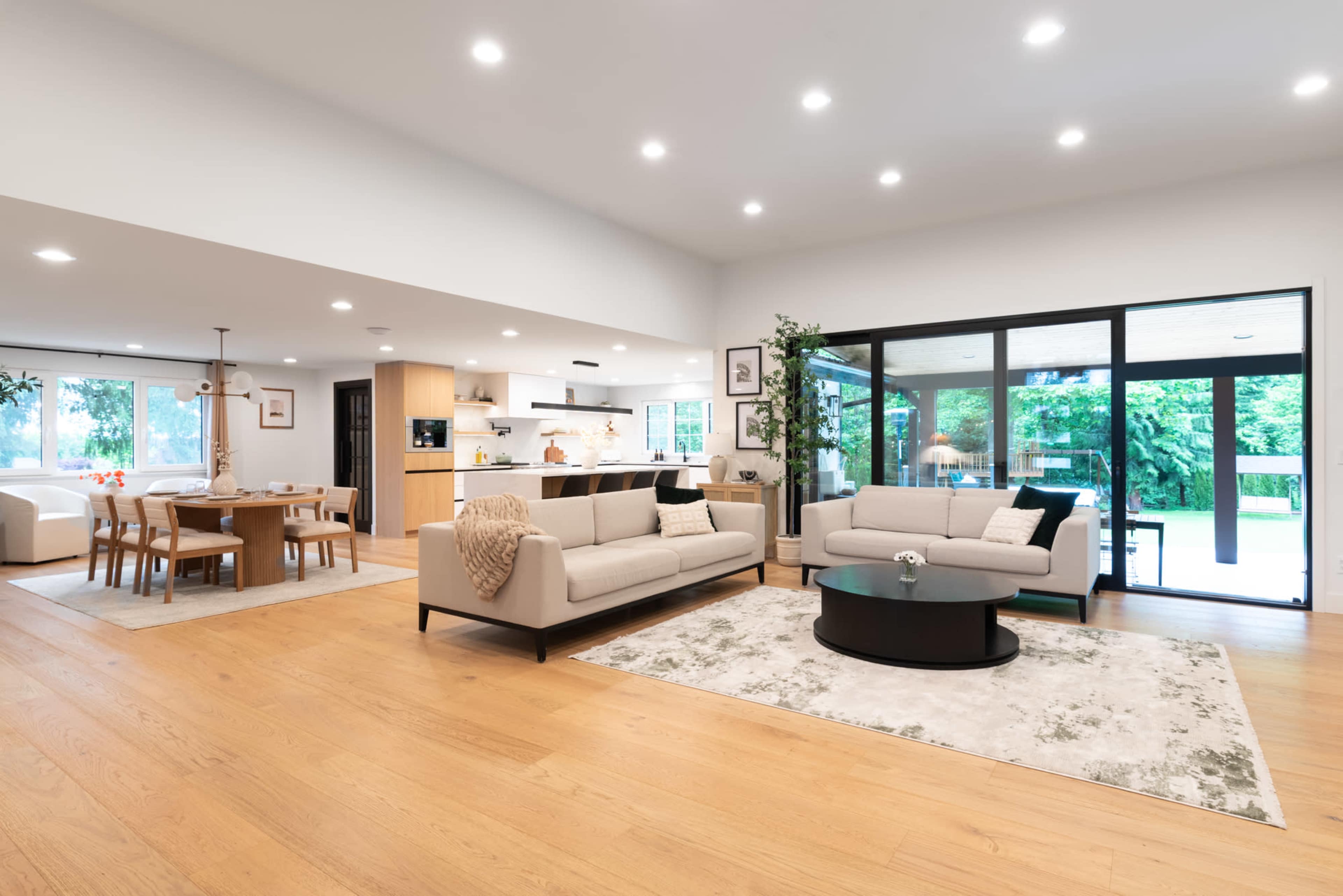 The image shows a modern living room with two sofas, a coffee table, and a dining area, all featuring large windows that open to a patio.