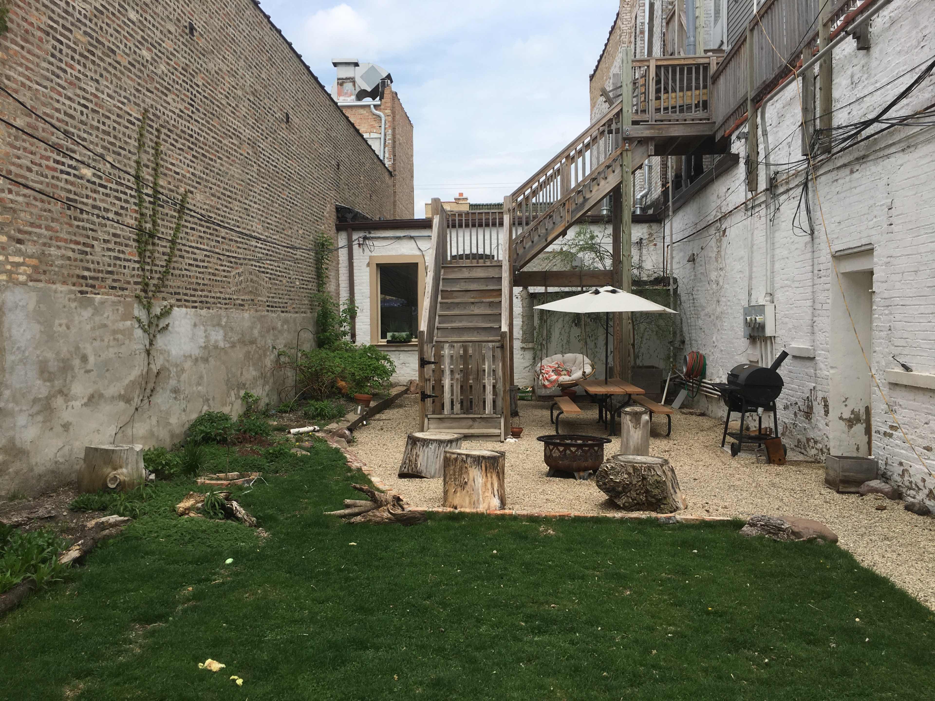 A gravel and grass backyard with a wooden deck, outdoor furniture, a fire pit, and a grill.
