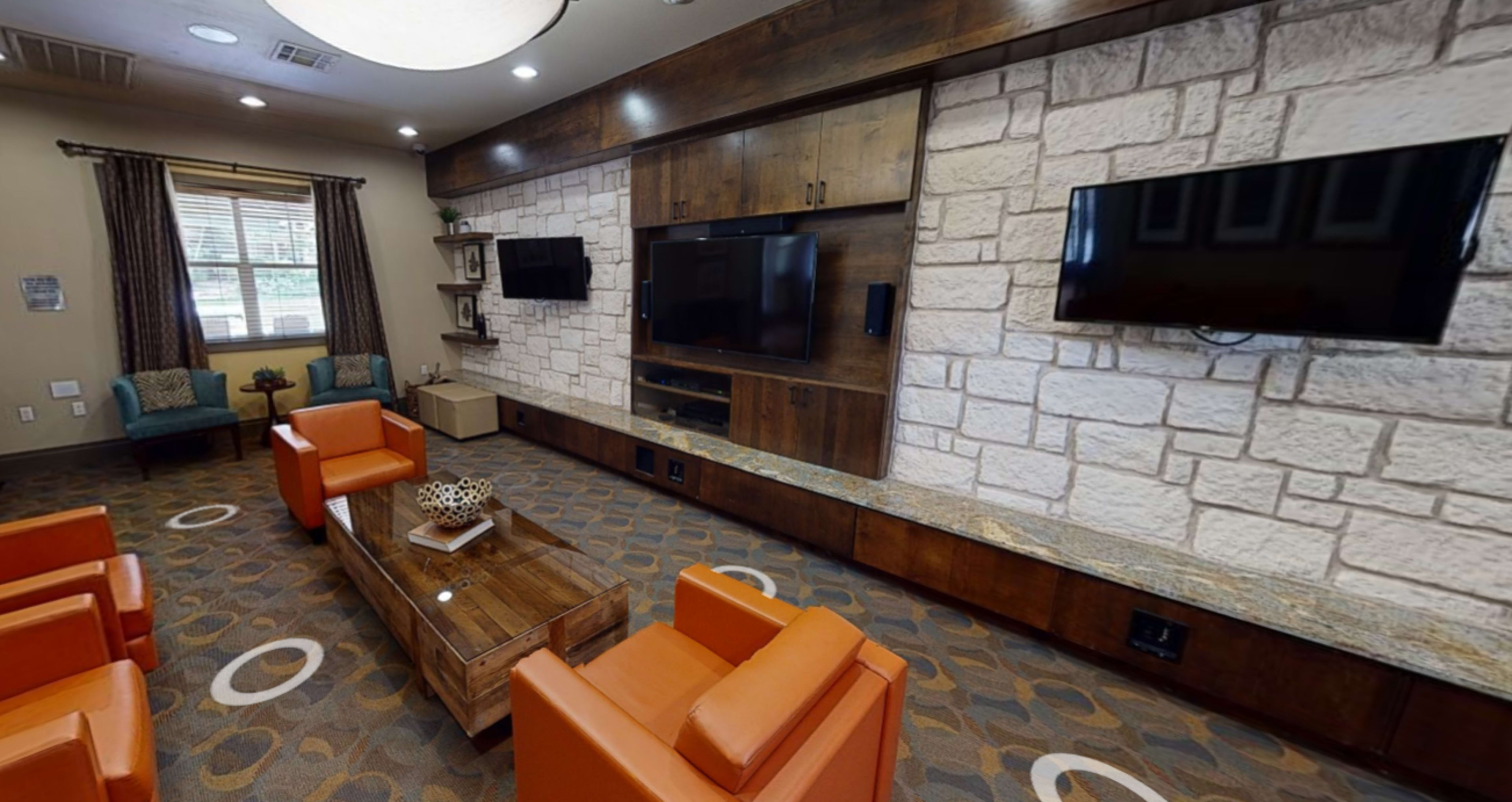 A lounge area with two orange chairs and a wooden coffee table, surrounded by a stone and wood feature wall with two mounted televisions.