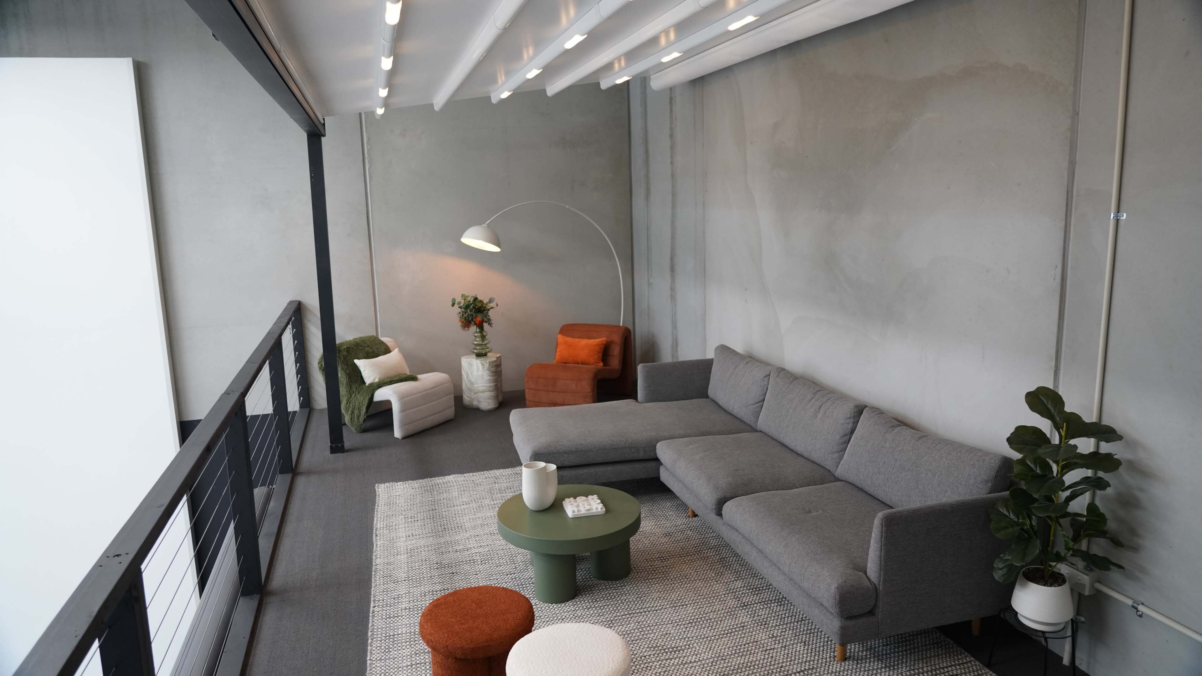 The image shows a modern, minimalistic living room with a gray sectional sofa, a green coffee table, and accent chairs in orange and white, set against a concrete wall.