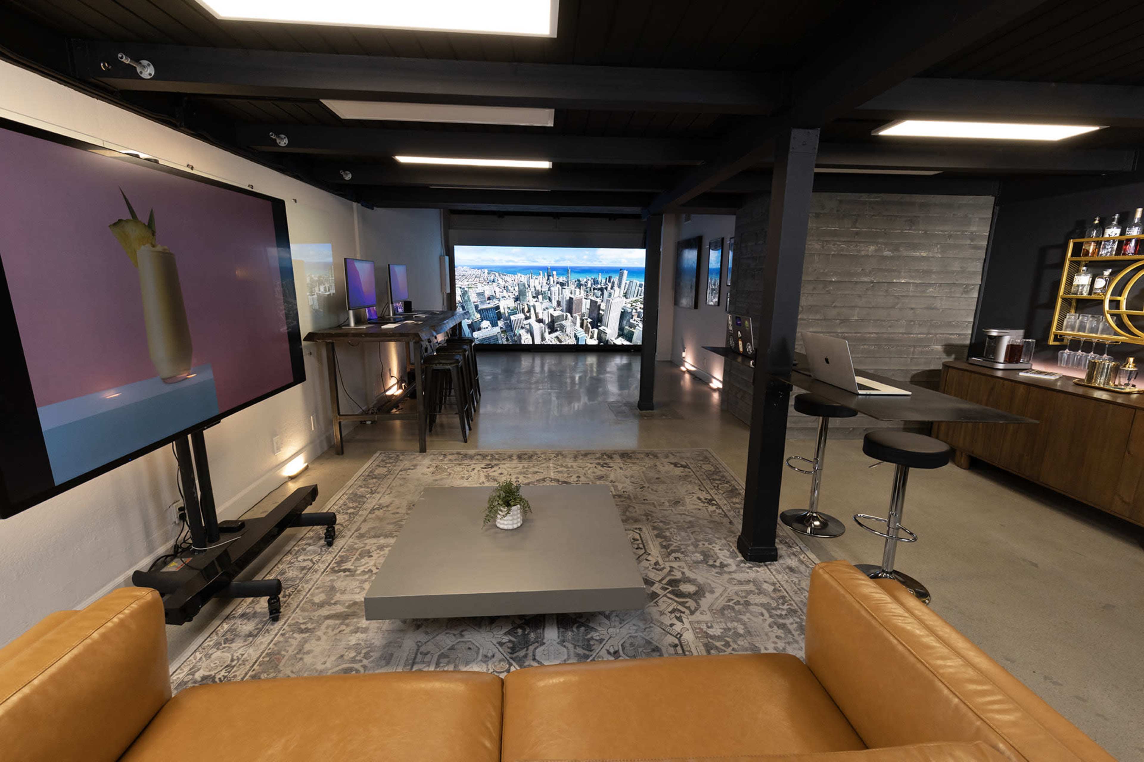 The image shows a modern interior space featuring a lounge area with a leather sofa, a large screen displaying a cityscape, and a bar counter with stools.