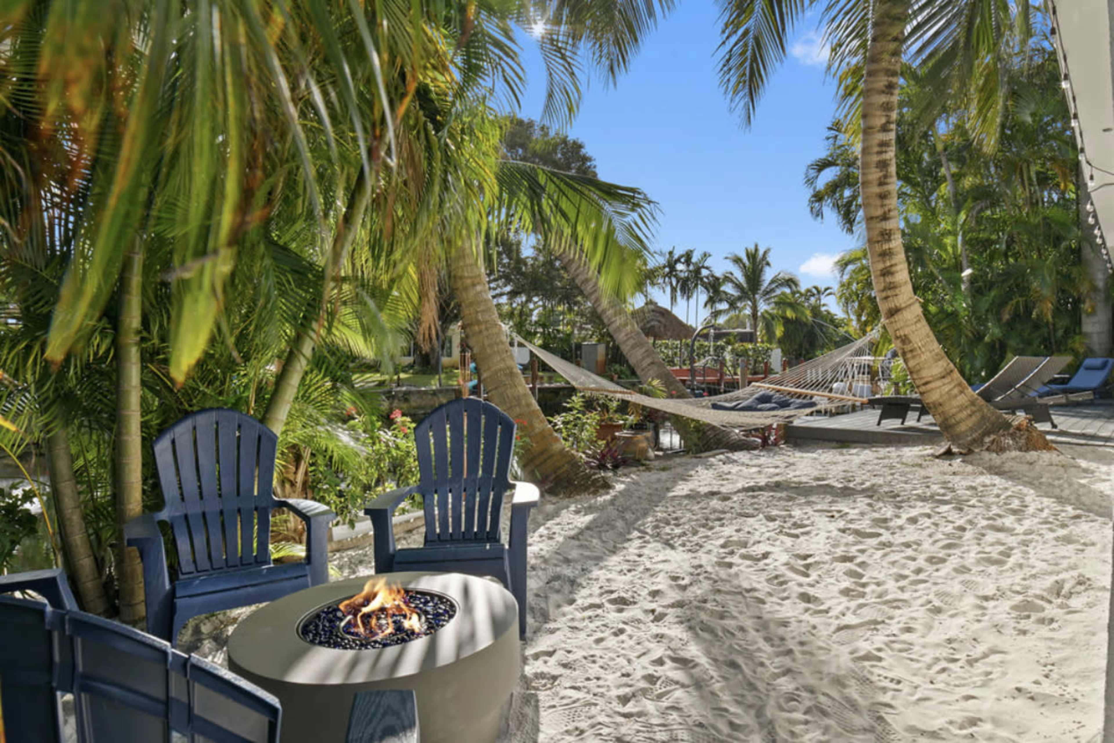 A sandy area features blue Adirondack chairs surrounding a fire pit, with palm trees and a hammock visible in the background.