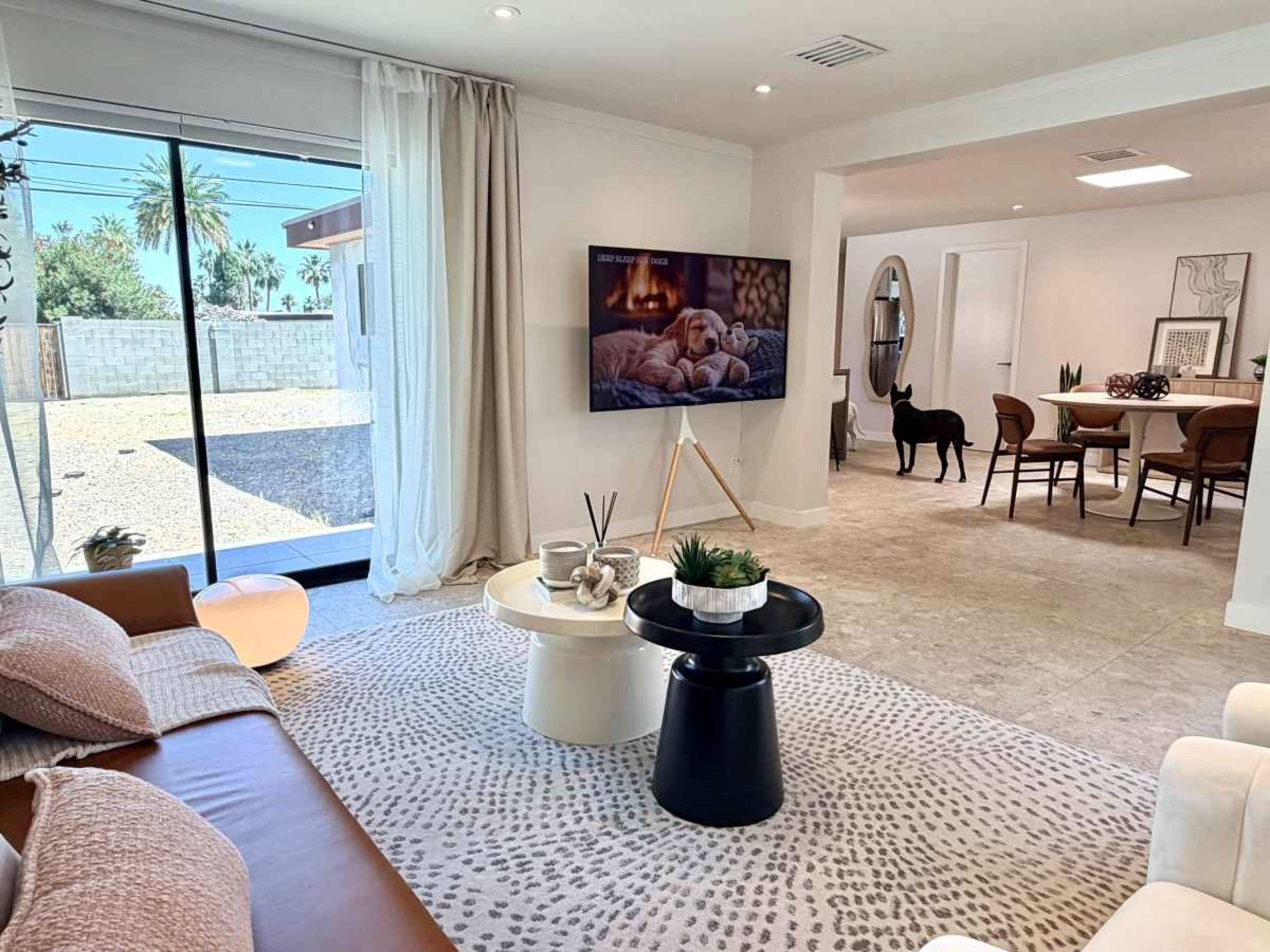 A modern, open-concept living area features a large TV displaying a dog, a sectional couch with decorative pillows, and a dining space in the background.