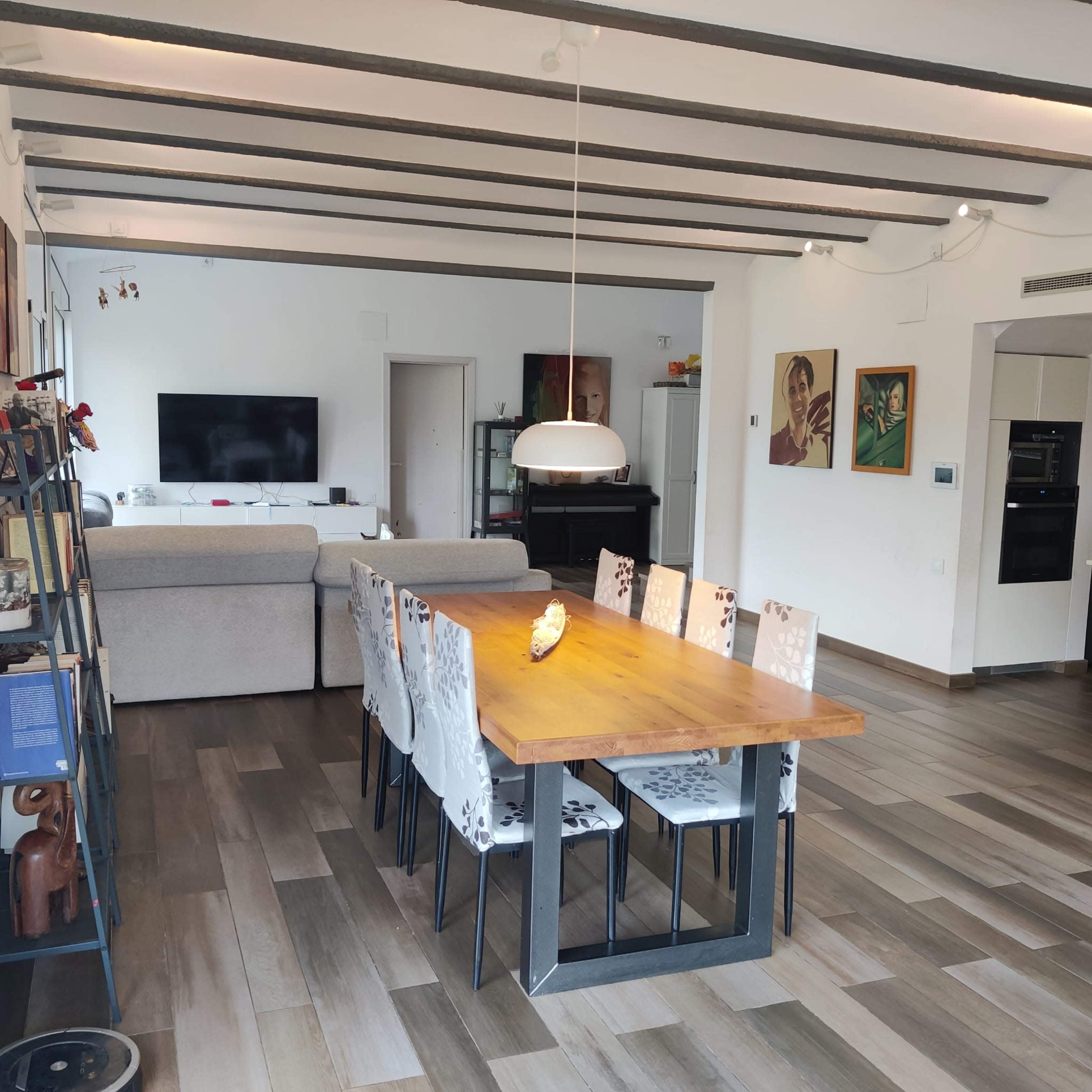 A modern living and dining area featuring a large wooden dining table with eight chairs, a gray sofa, and a television mounted on the wall.