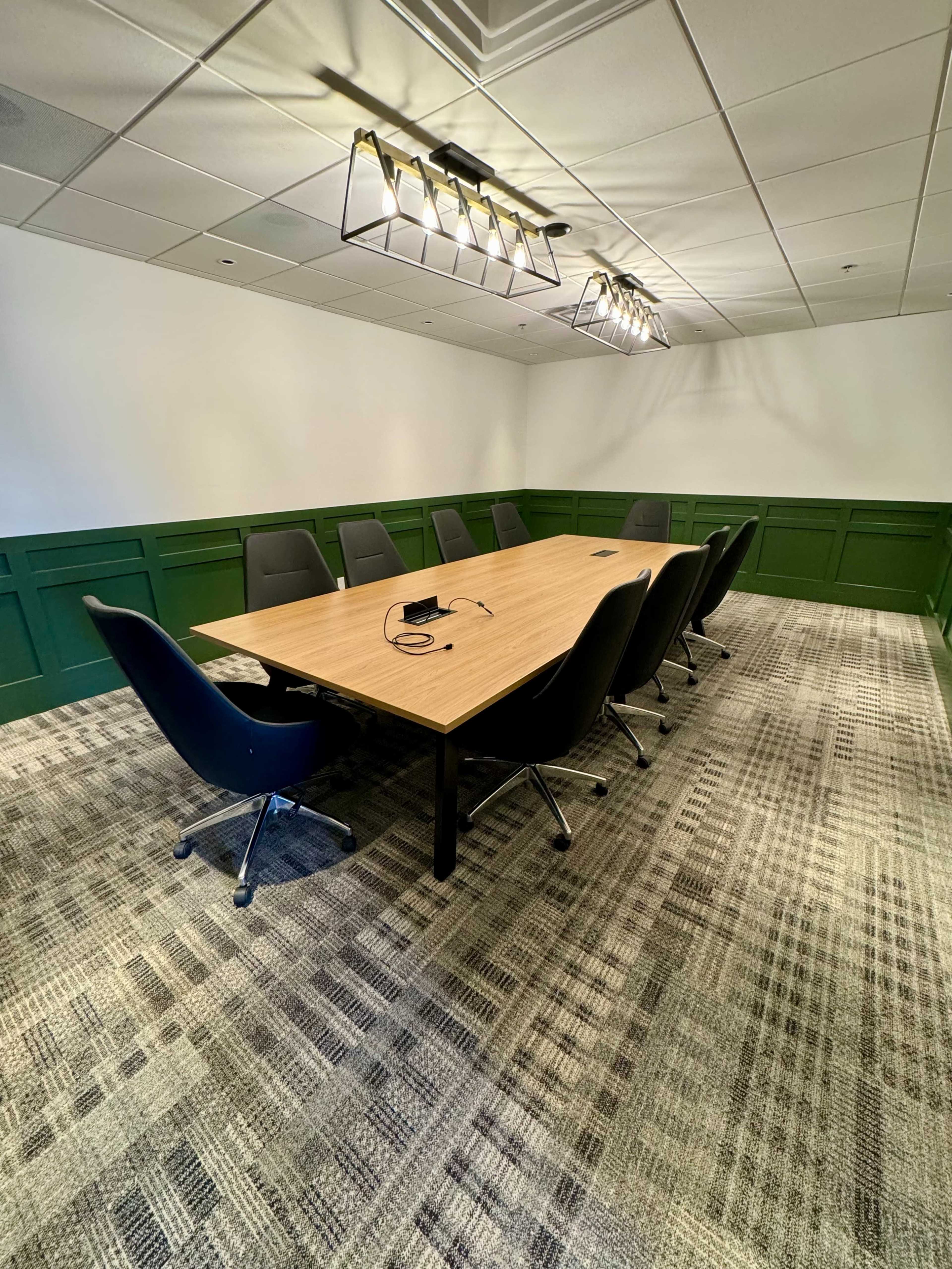 A conference room features a large wooden table surrounded by eight chairs and serves as a meeting space with green paneling and overhead lighting.