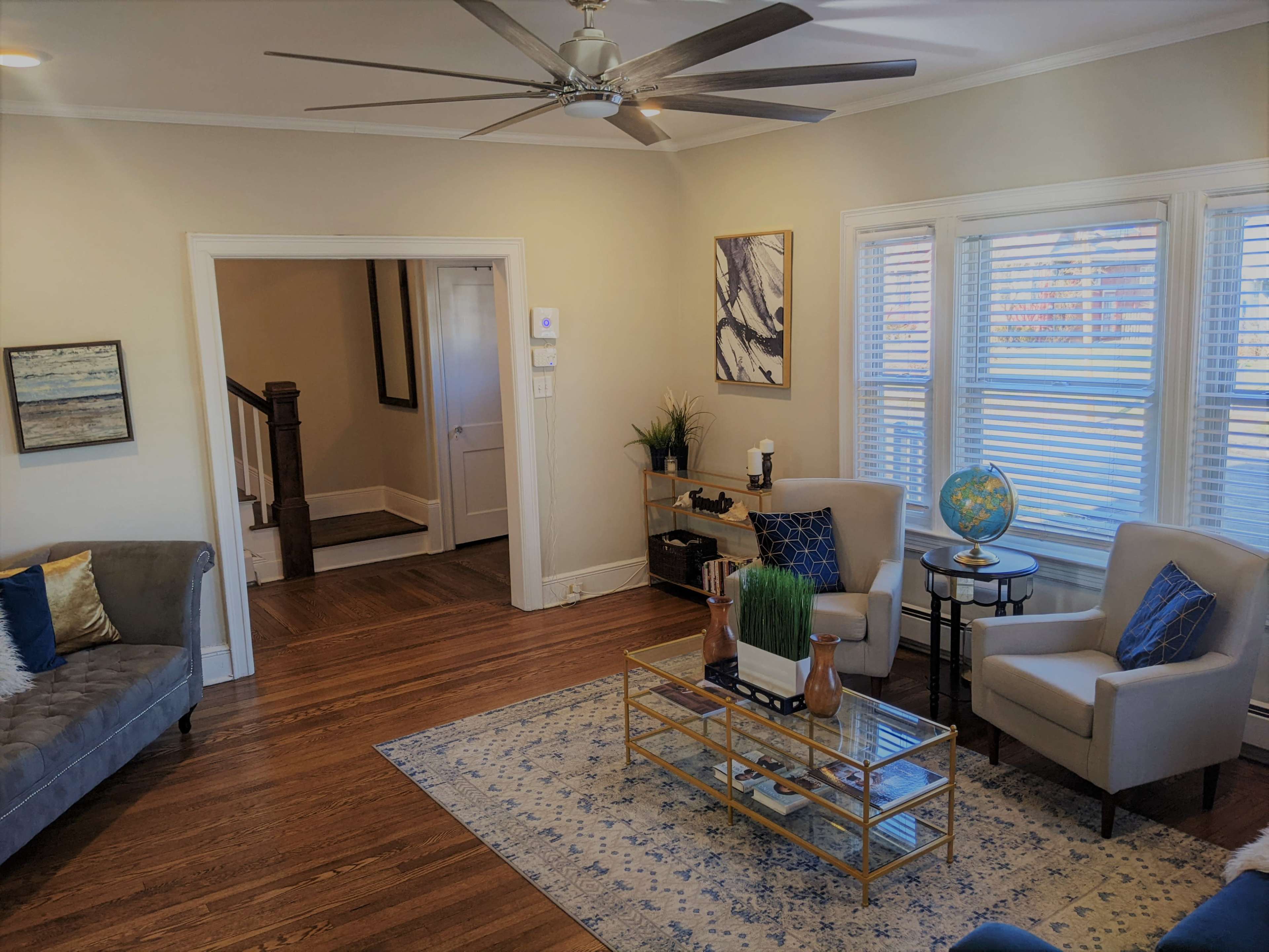 The image shows a living room with hardwood floors, a gray couch, two armchairs, a coffee table, and a staircase leading to another area.